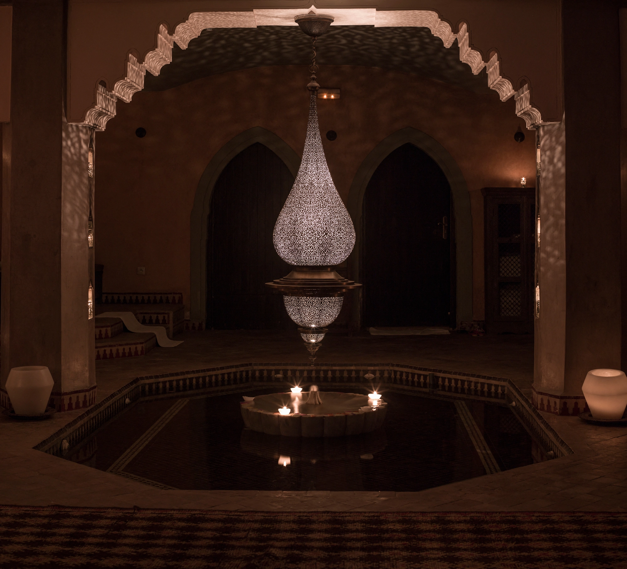 A pierced teardrop pendant lamp glowing in an arched dark room above a reflecting pool.