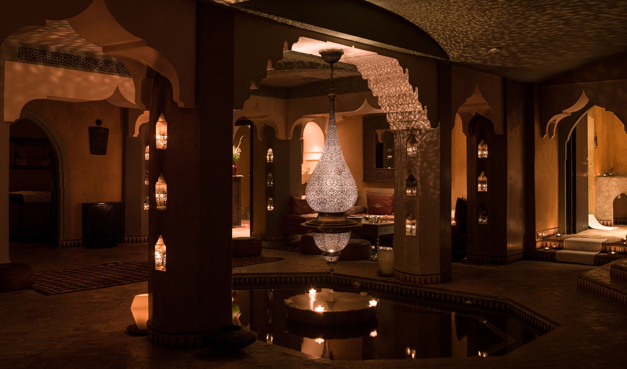 Dim warm-lit hammam with arched alcoves and lanterns reflected in a still indoor pool.