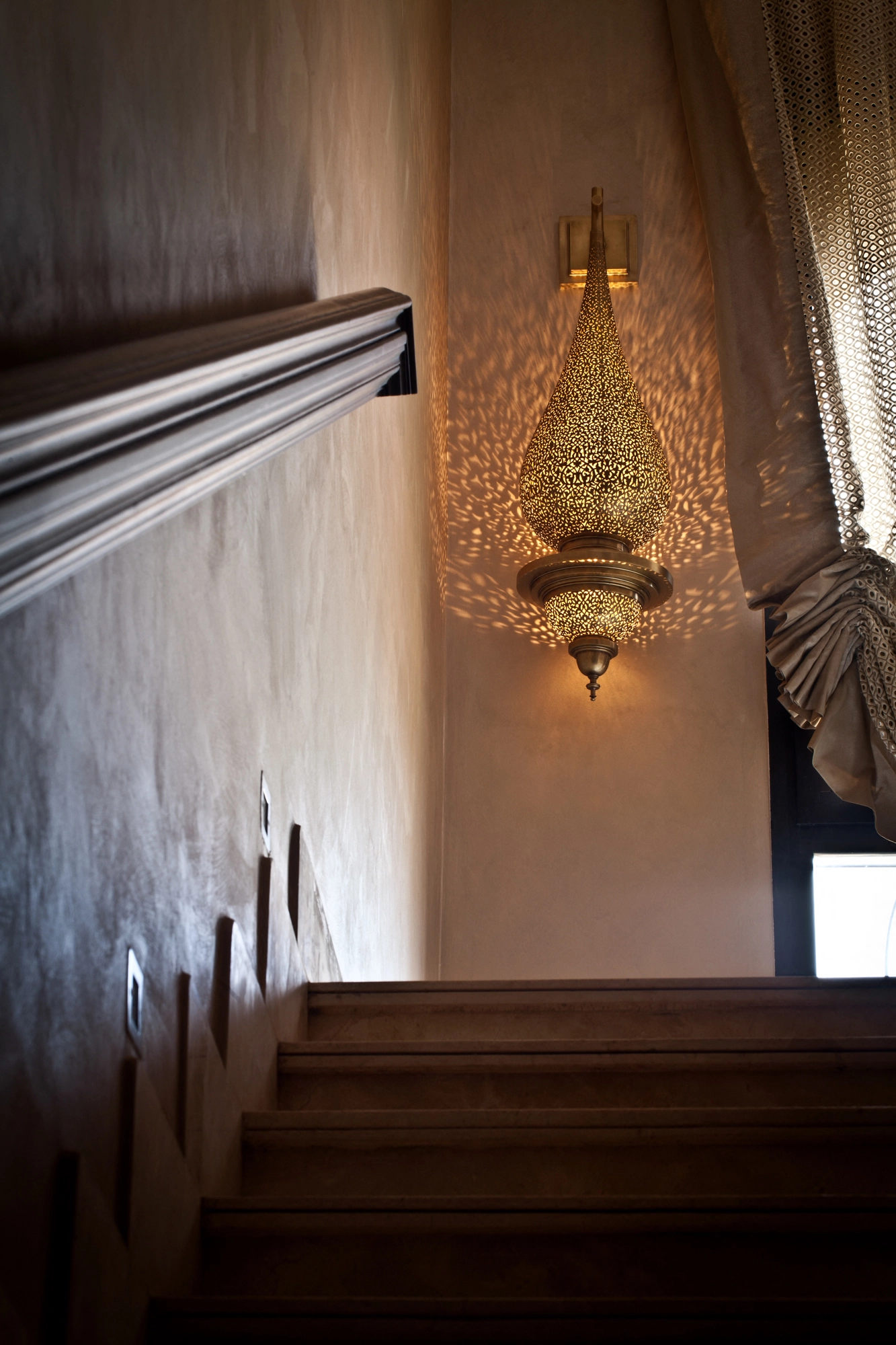 A pierced teardrop pendant lamp glowing warmly in a stairwell beside a draped curtain.