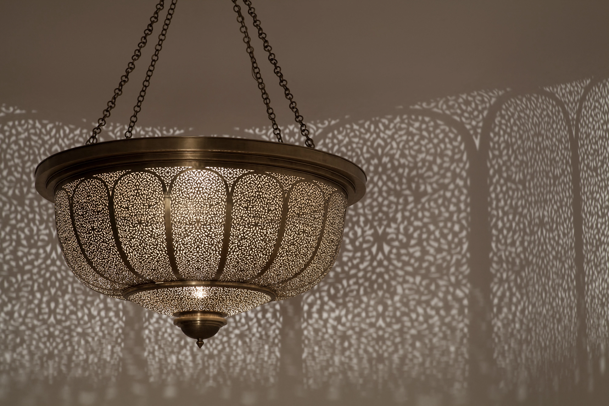 Brass pierced bowl-shaped Nafura pendant hung on chains, glowing against a wall flecked with light dots.