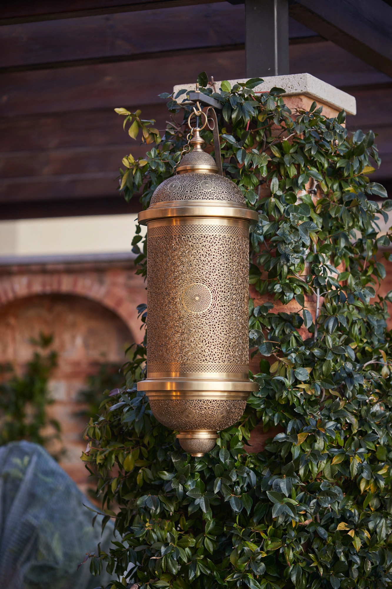 Brass pierced cylinder pendant lantern hanging outdoors beside a brick arch and climbing greenery.