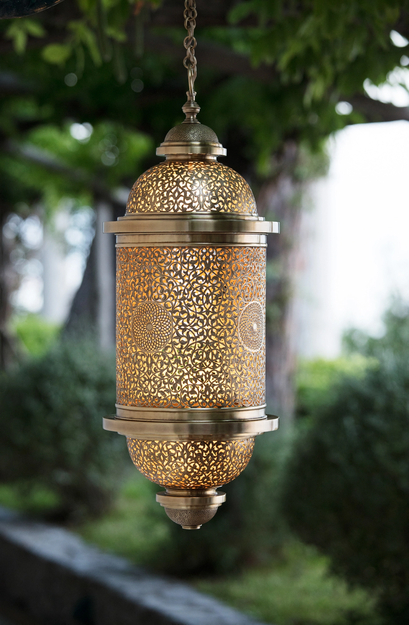 Brass pierced cylinder pendant lantern hanging outdoors against a clipped green hedge.