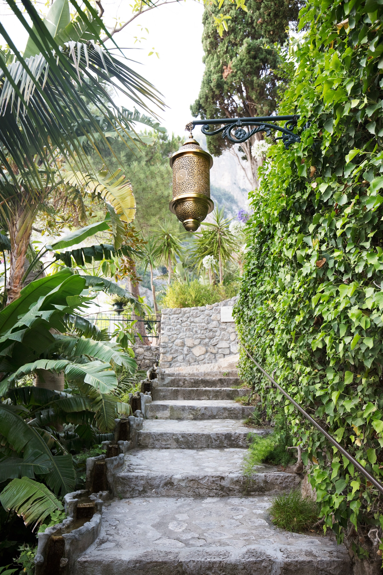 Brass cylinder pendant lantern hanging above a garden stone staircase framed by lush greenery.