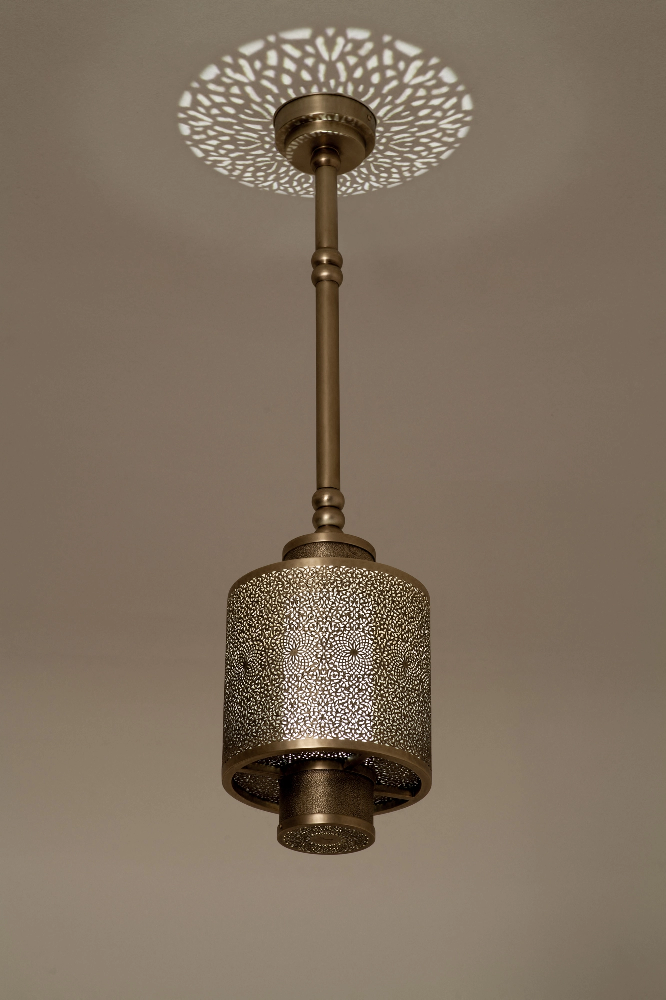Pierced-brass cylinder pendant on a slim stem, glowing beneath a floral rosette of projected ceiling light.