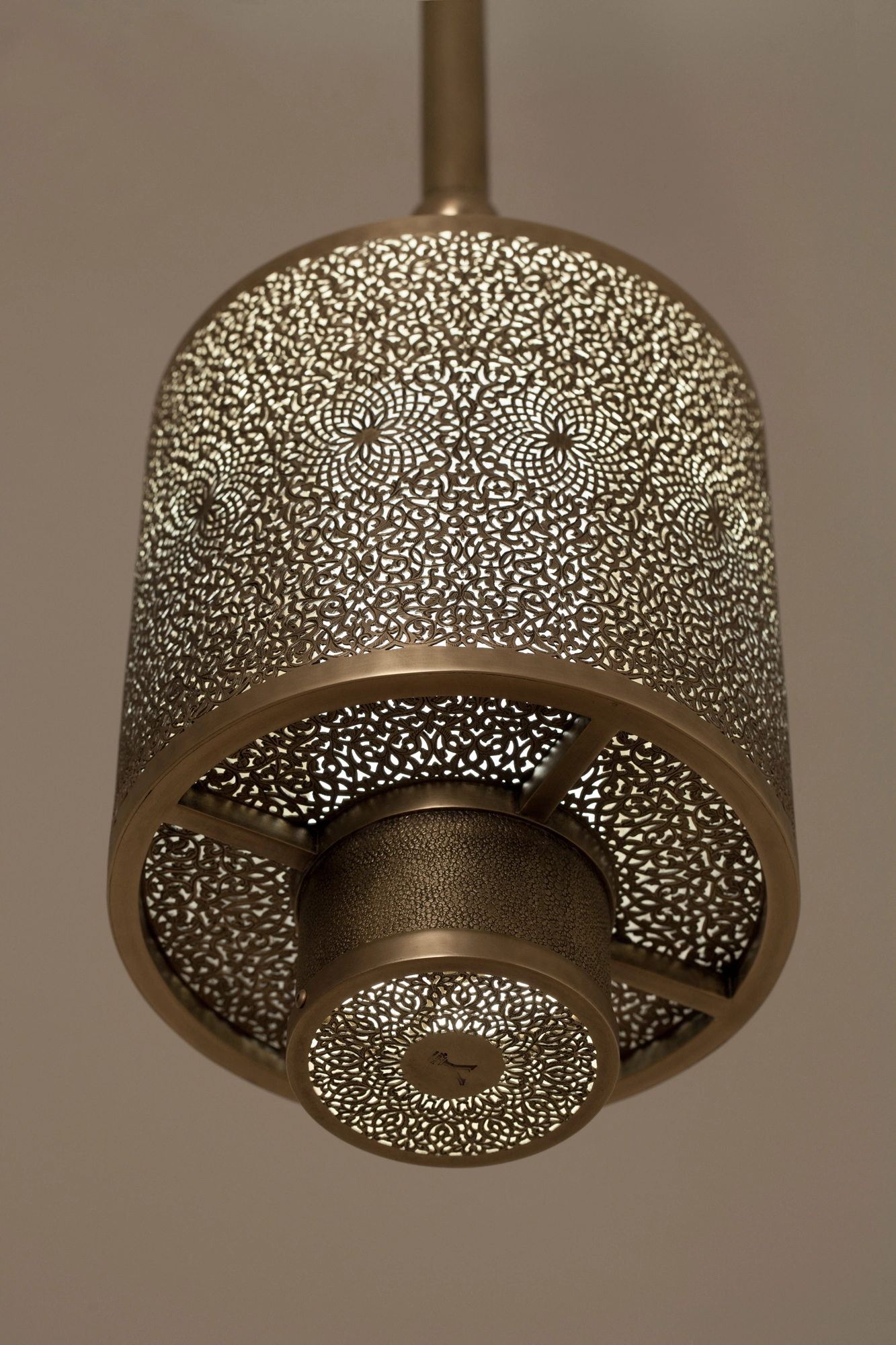 Double-walled pierced-brass cylinder pendant suspended from a stem, glowing warmly against a beige wall.