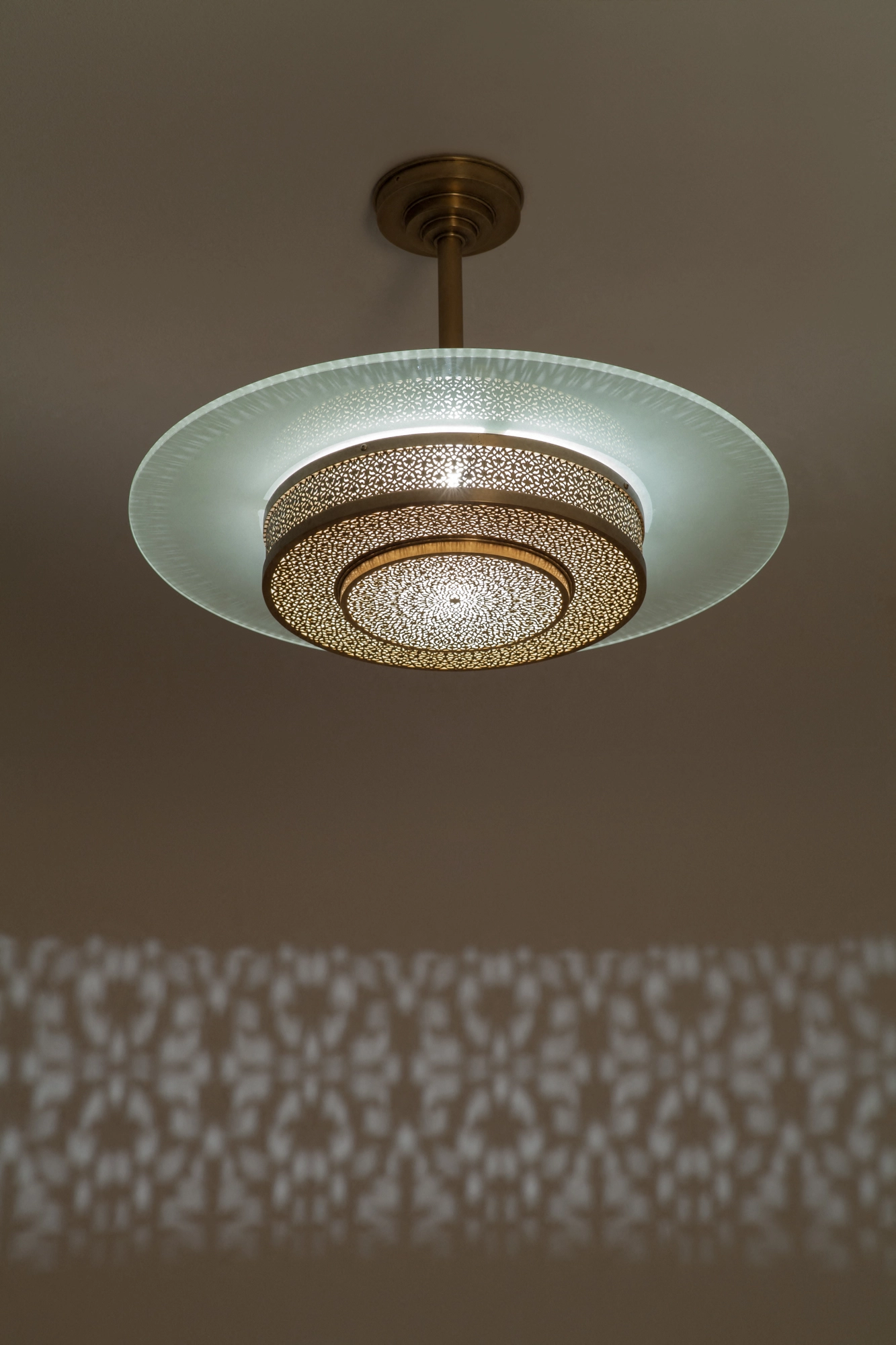Tiered frosted-glass disc-drum pendant with a pierced-brass band, glowing softly beneath a patterned ceiling.