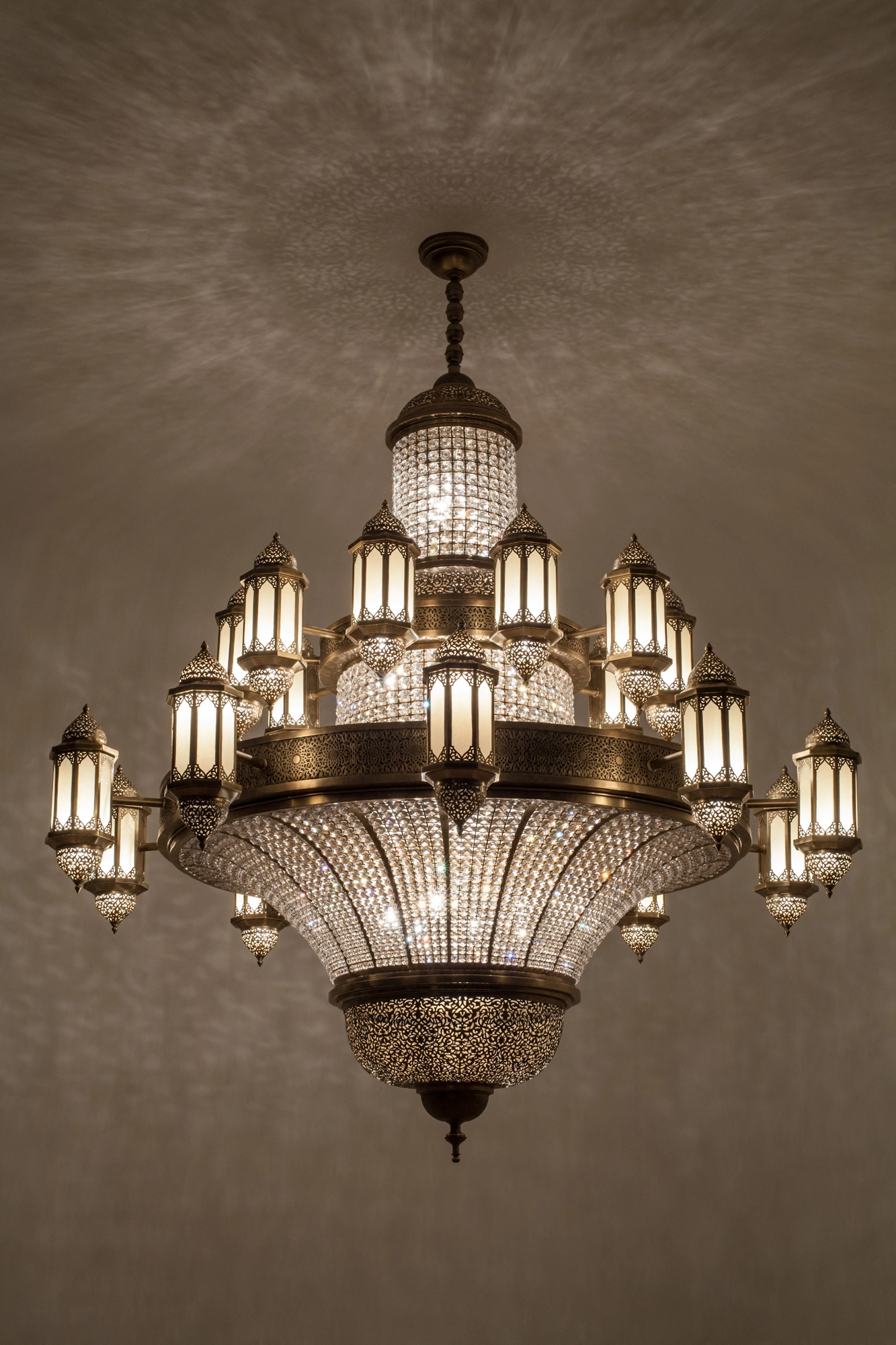 A large multi-tier chandelier of crystal beading and pierced brass lanterns, glowing warmly against a pale wall.
