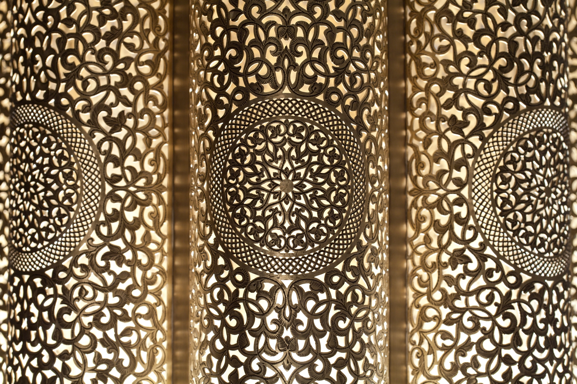 Close-up detail of a pierced-brass lantern surface, its floral perforations glowing warm gold against shadow.