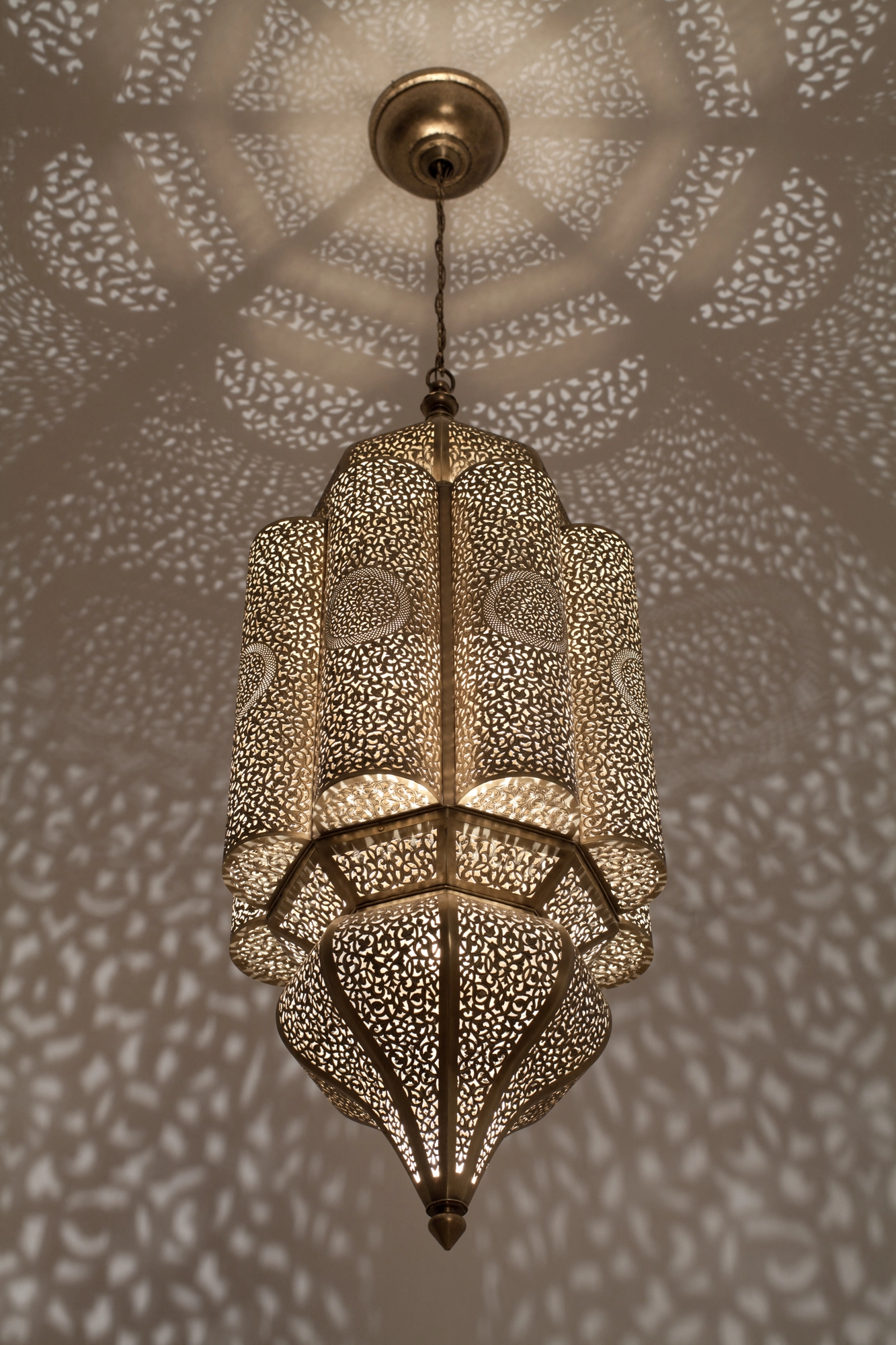 Pierced-brass Andalous pendant seen from below, its flared tiers glowing amid a soft scatter of light on the ceiling.