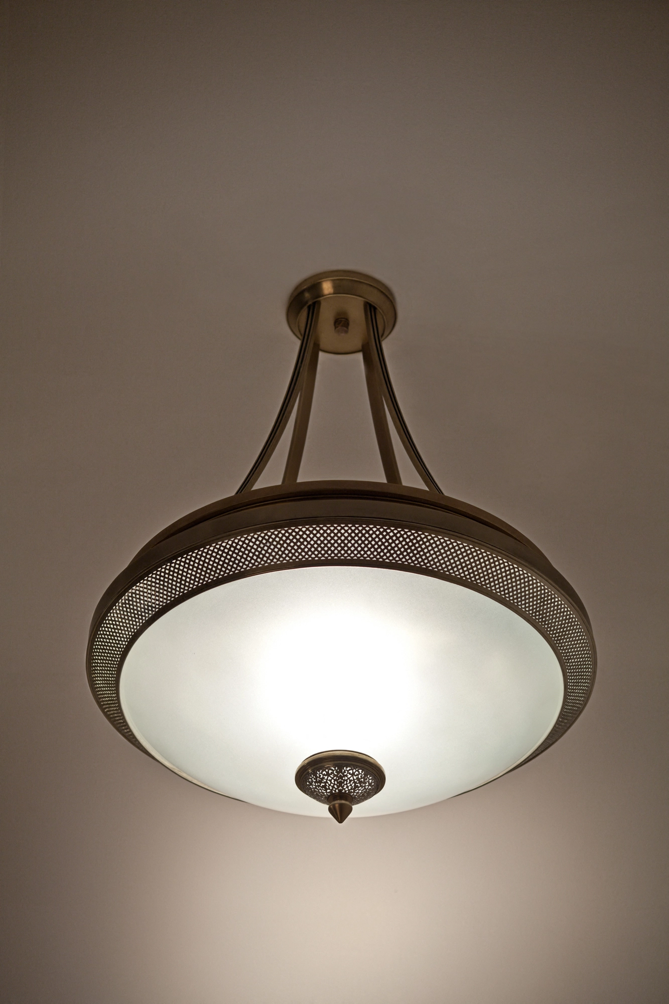 Frosted-glass bowl pendant cradled in a pierced-brass ring on three arms, glowing warmly against a pale ceiling.