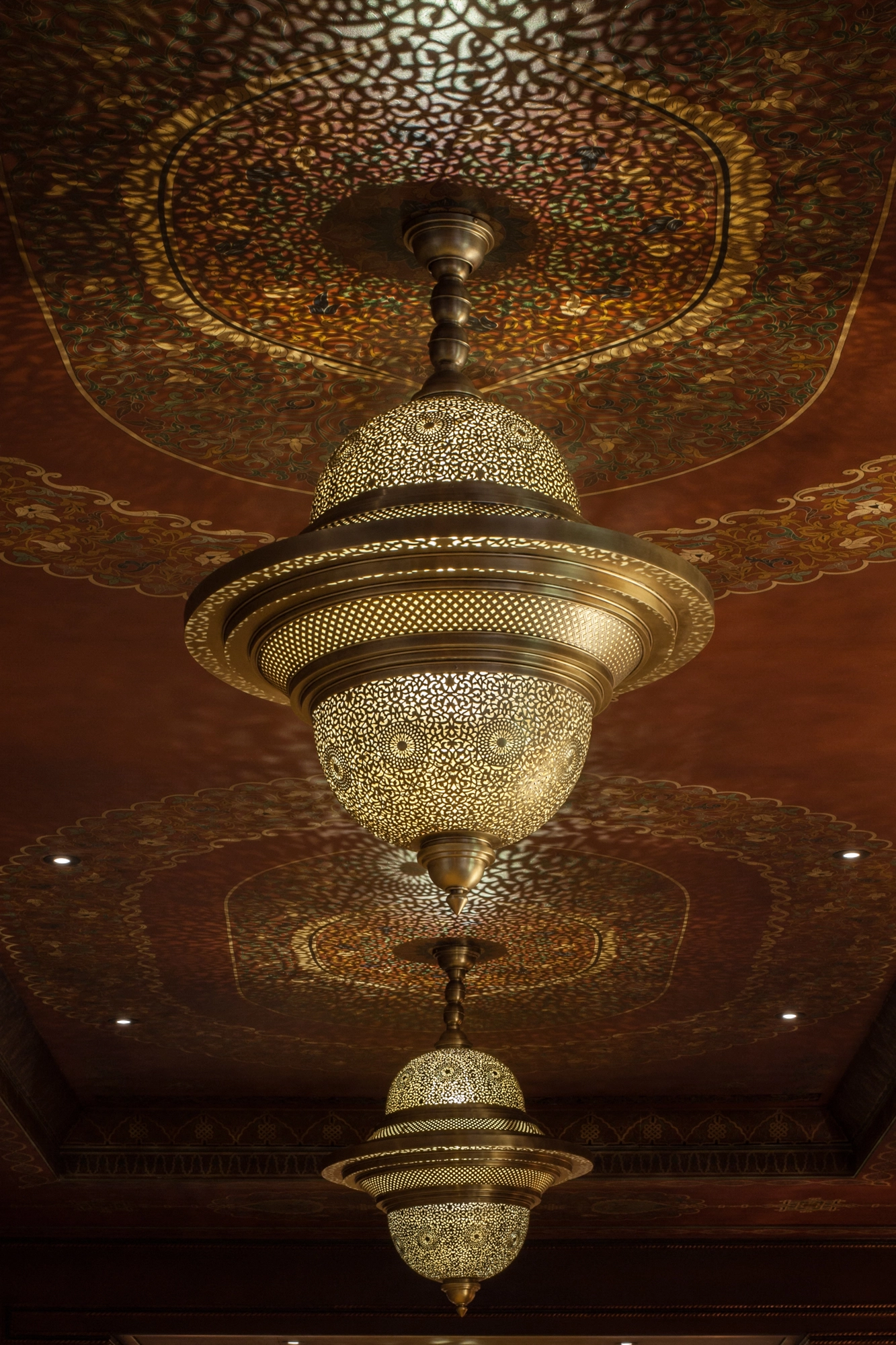 Pierced twin-dome brass pendant hanging from an ornate gilded ceiling, glowing warmly in a dim room.