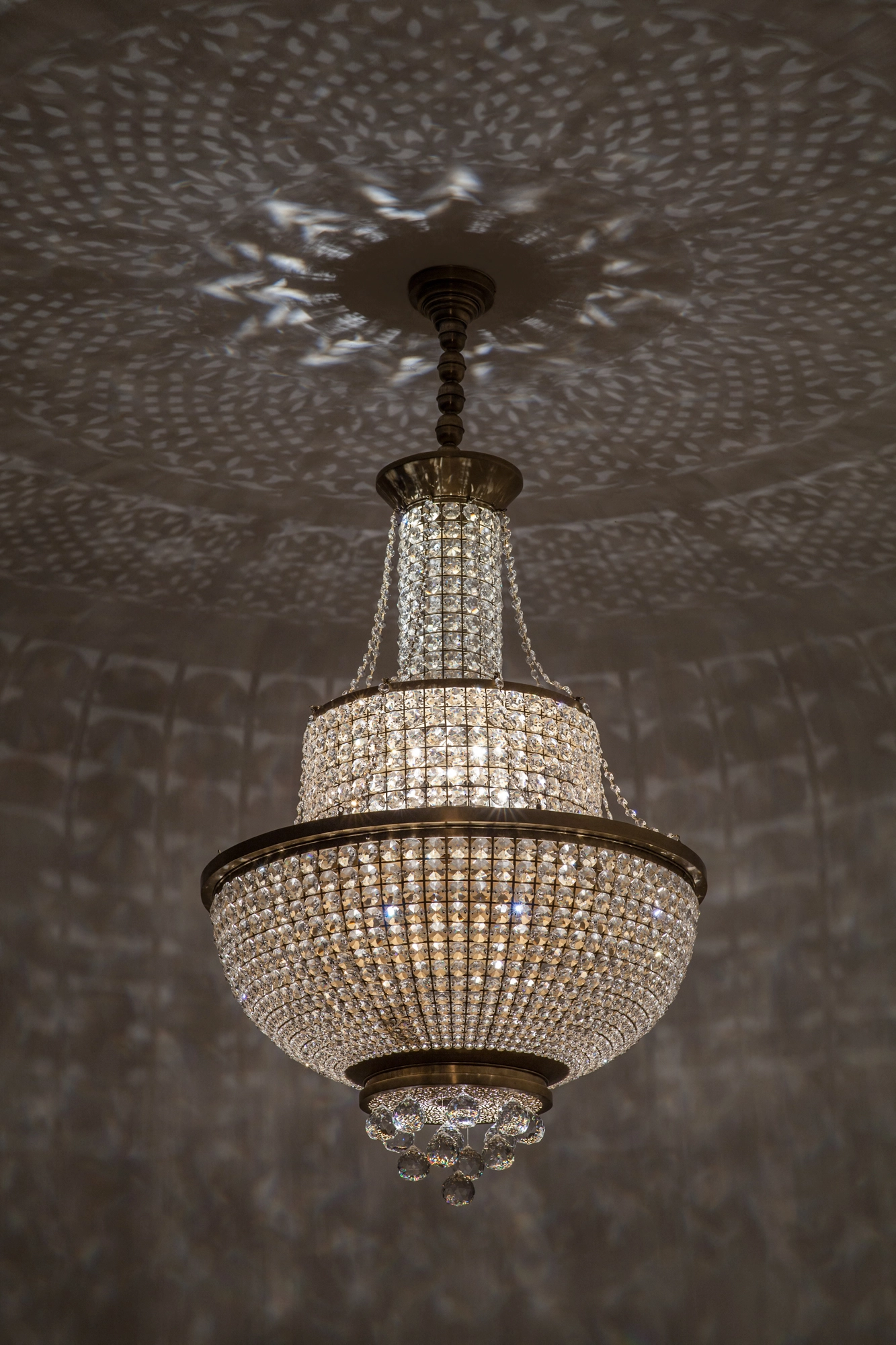 A three-tier crystal basket chandelier suspended from a dark ceiling scattered with dappled light.