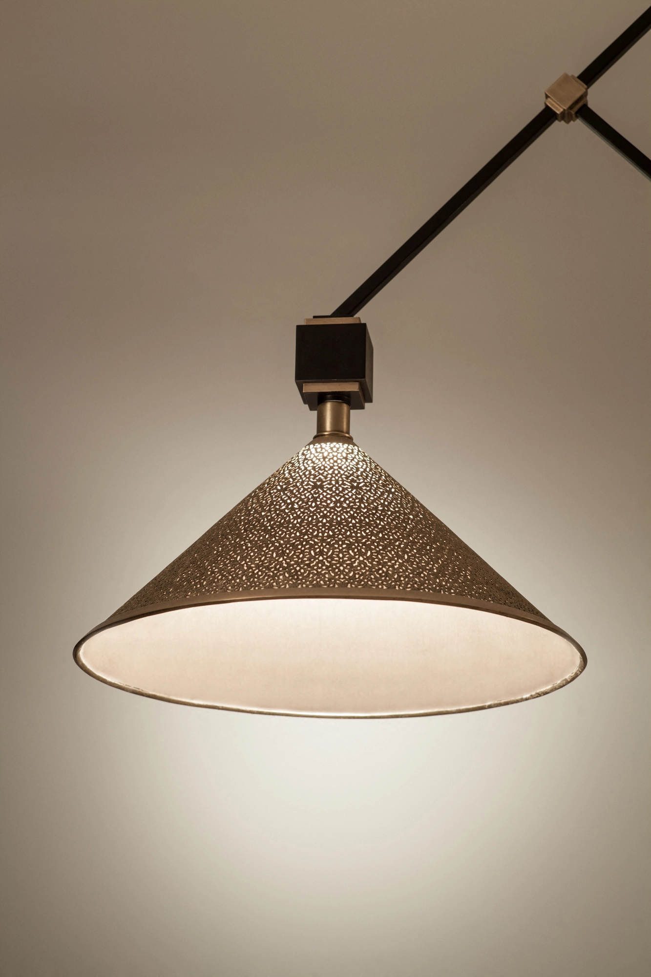 A single conical pendant shade glowing warmly from below, suspended on a dark rod against a pale background.