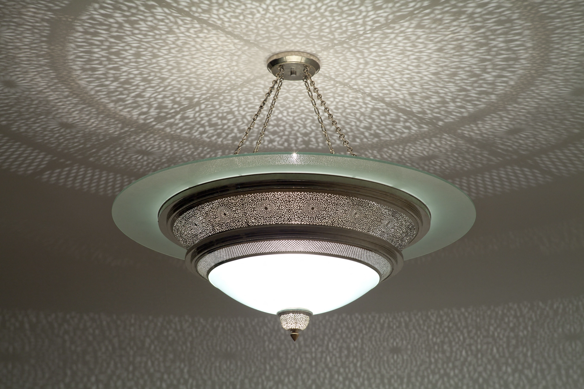 Frosted glass disc-dome ceiling light with a pierced-brass band, set against a ceiling of soft light patterning.