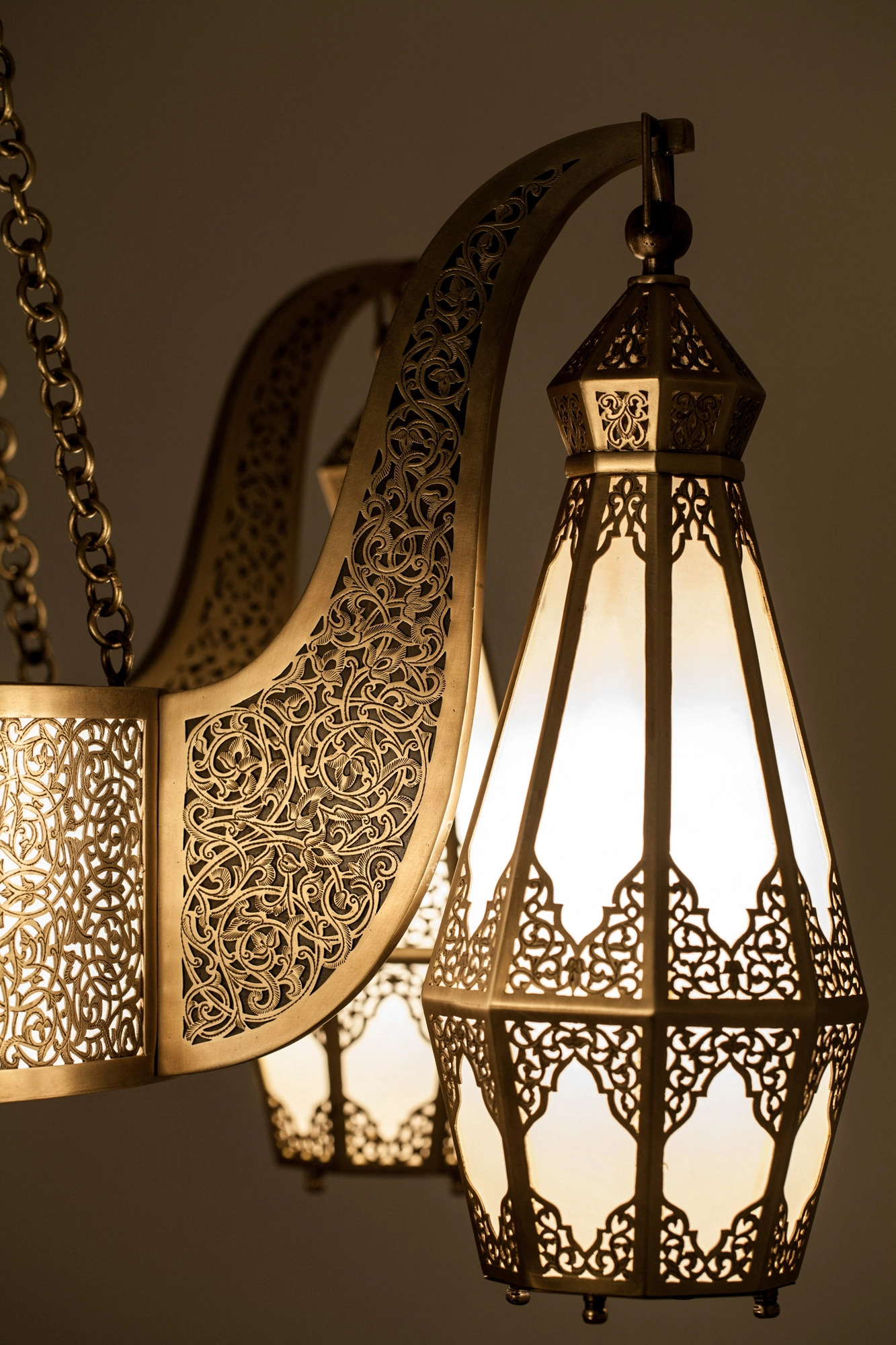 Close-up of a pierced brass lantern shade and a scrolled chandelier arm, lit warmly against a dim wall.