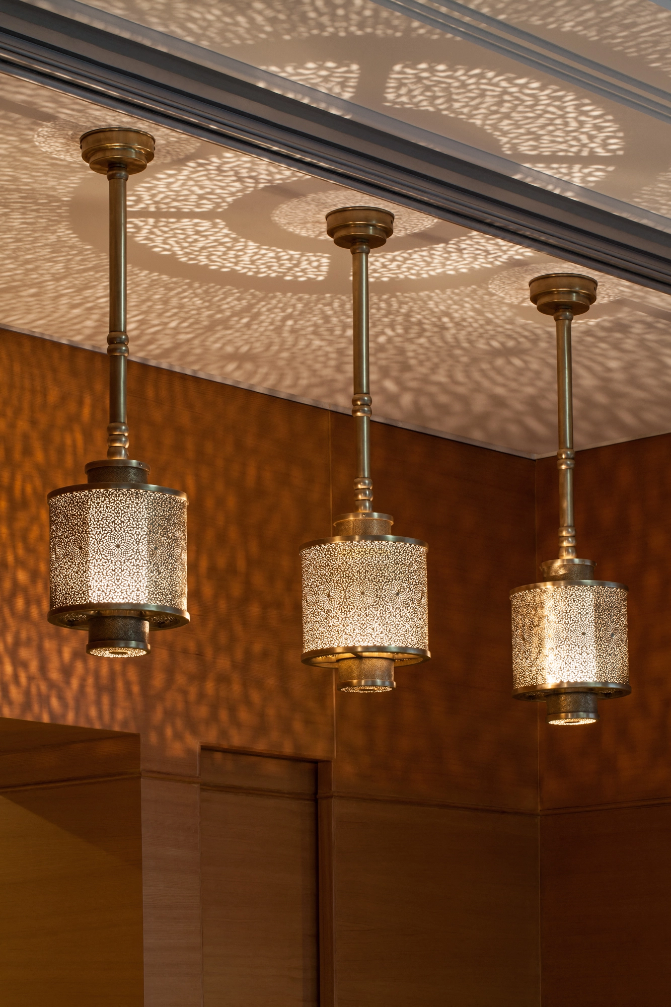 Three pierced cylinder pendant lanterns hanging in a row in a warm orange-toned interior.