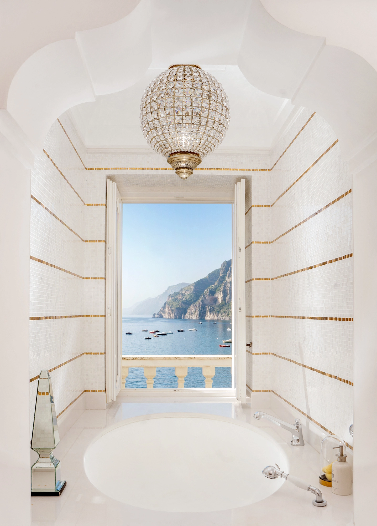 Crystal globe pendant above a bathtub set in a white niche framing a sea view through the window.