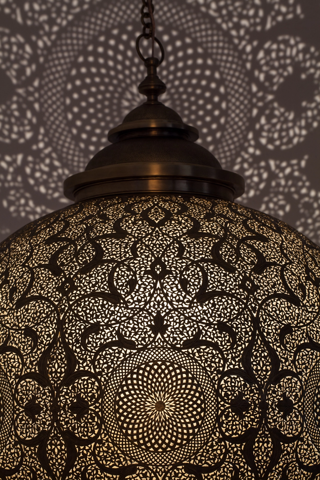 Close-up of a pierced-brass globe lantern's lower half, its dense floral perforations glowing gold against black.