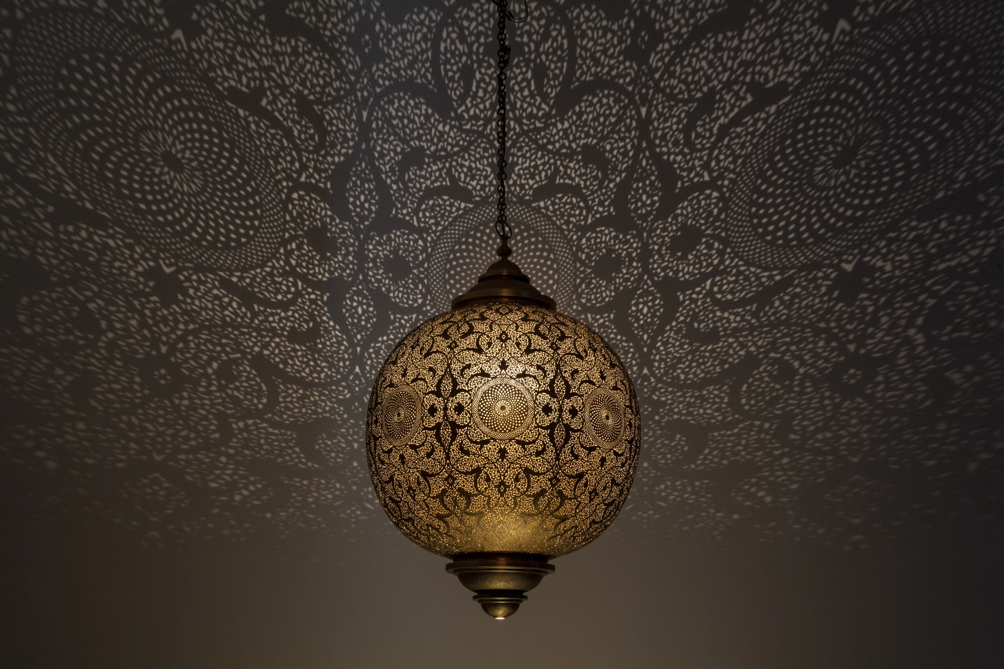Pierced-brass globe pendant with a tiered hat, seen from below and glowing amid scattered ceiling light.