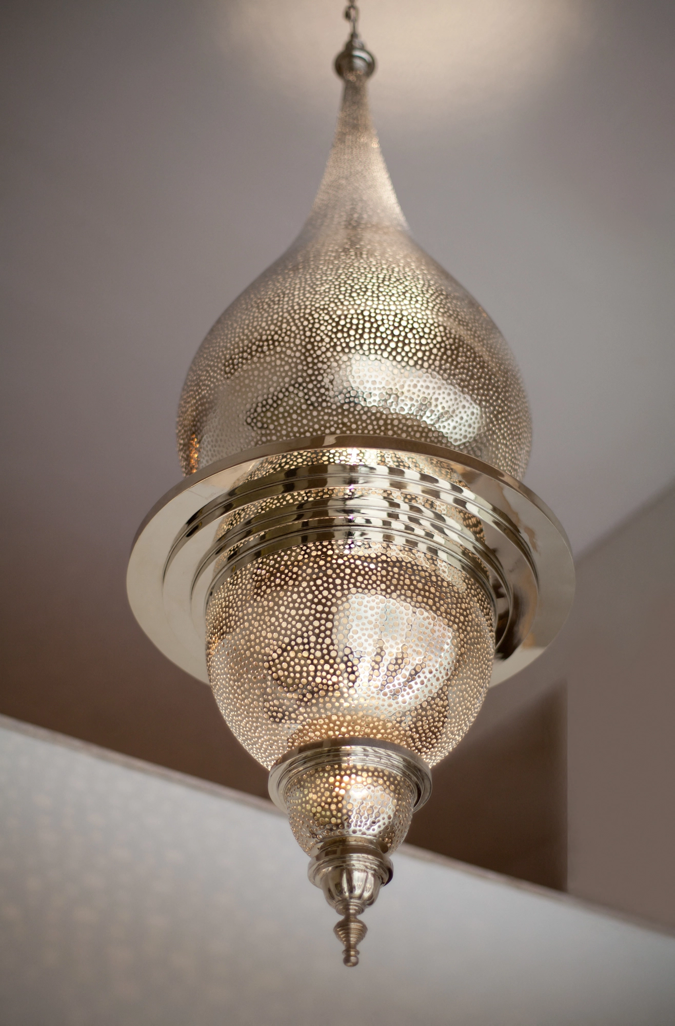 Silver pierced onion-domed pendant lamp with a pointed finial, glowing against a pale wall corner.