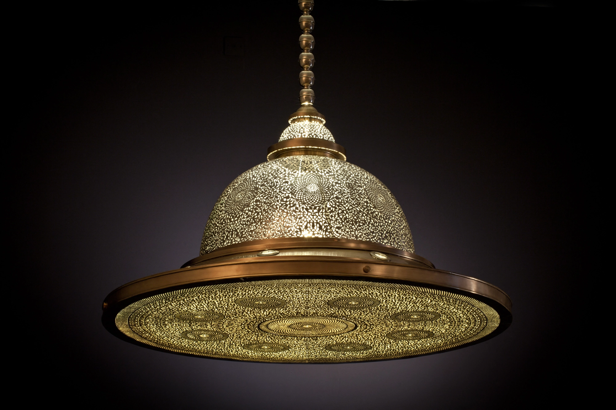 Glowing pierced-brass domed medallion pendant with a flat disc brim, suspended by a chain against black.