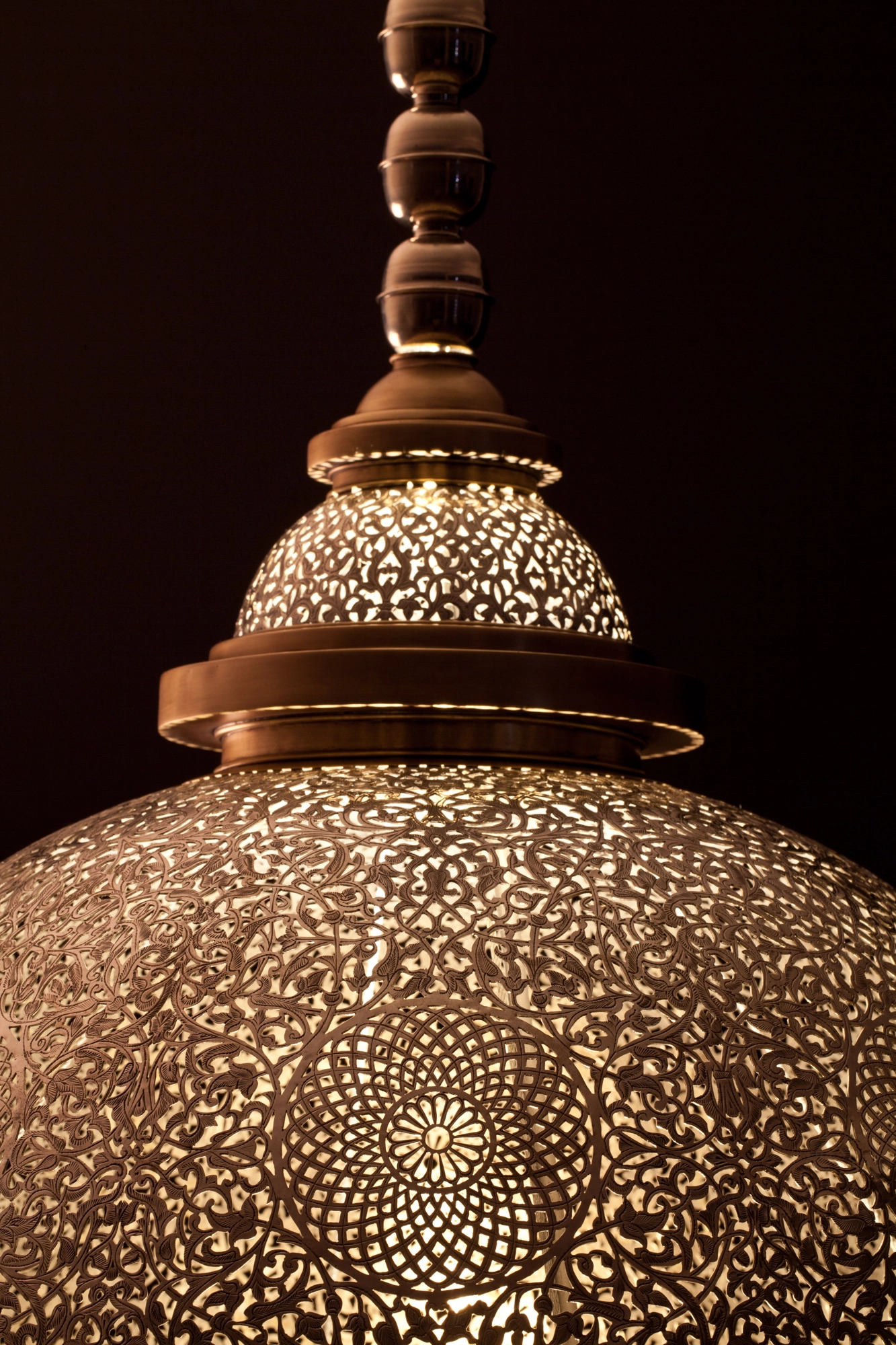 Underside of a pierced-brass domed medallion pendant, its radiating floral perforations glowing warm gold.