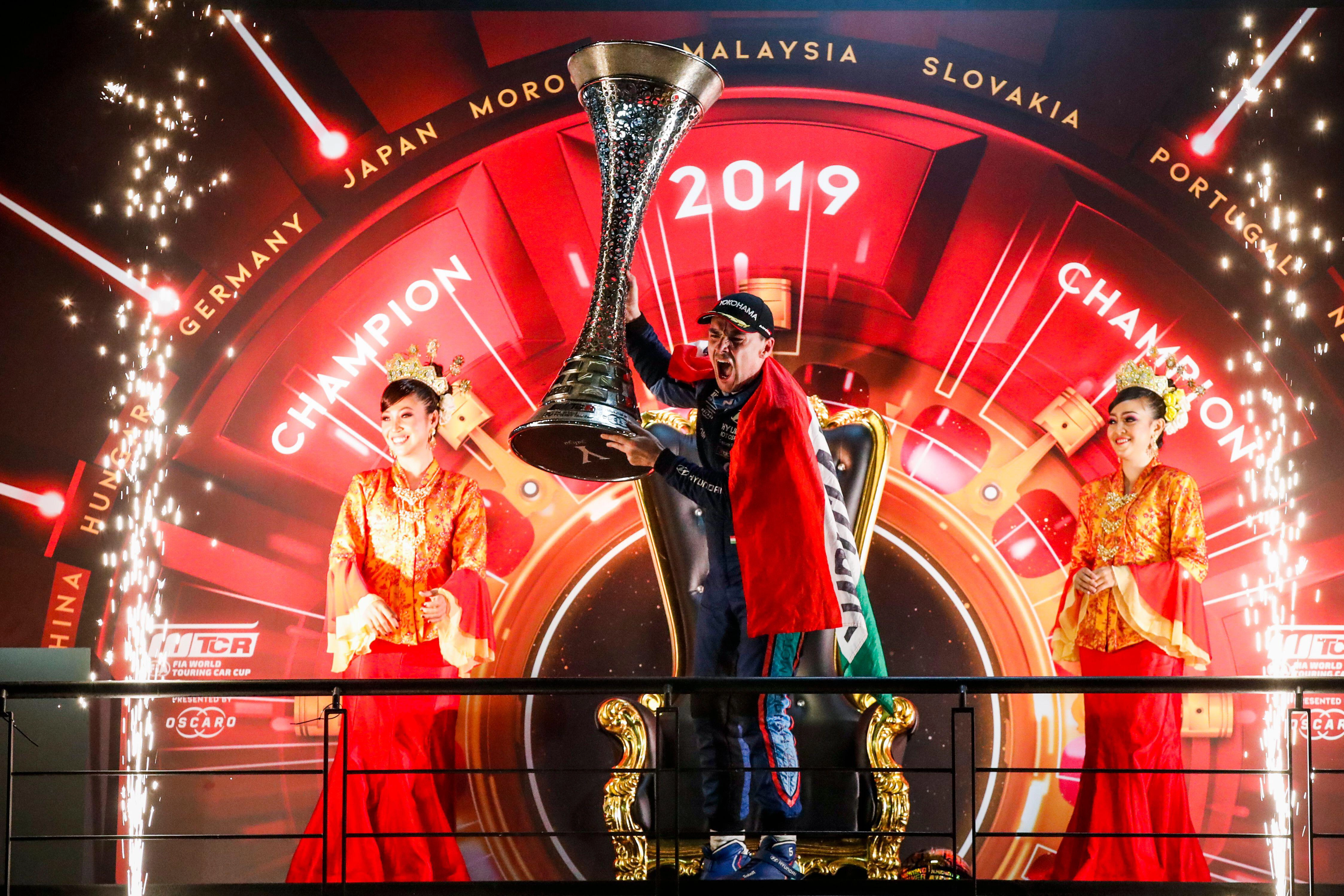 A driver celebrates on a fireworks-lit podium, lifting a trophy before a 2019 championship backdrop.