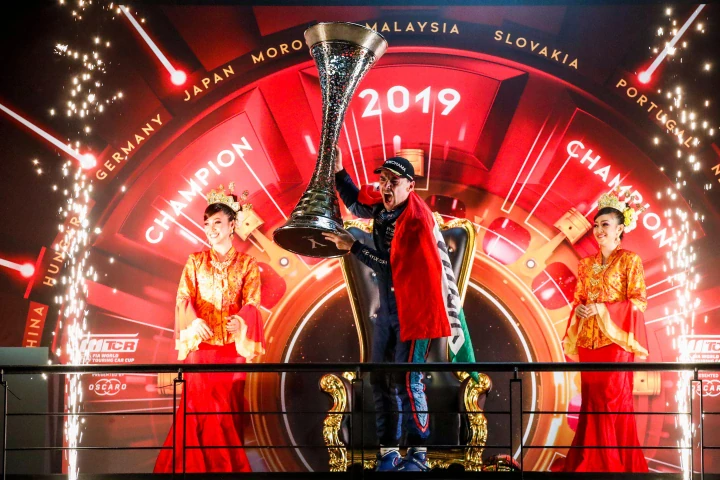 A driver celebrates on a fireworks-lit podium, lifting a trophy before a 2019 championship backdrop.
