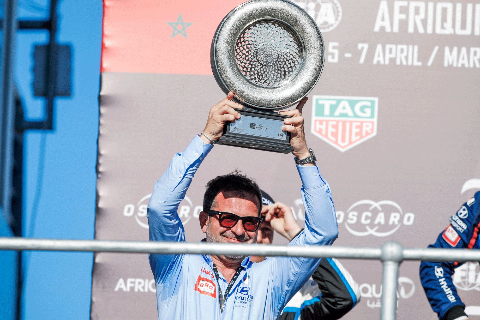 A person holds the pierced trophy plate aloft at a race event in front of event banners.