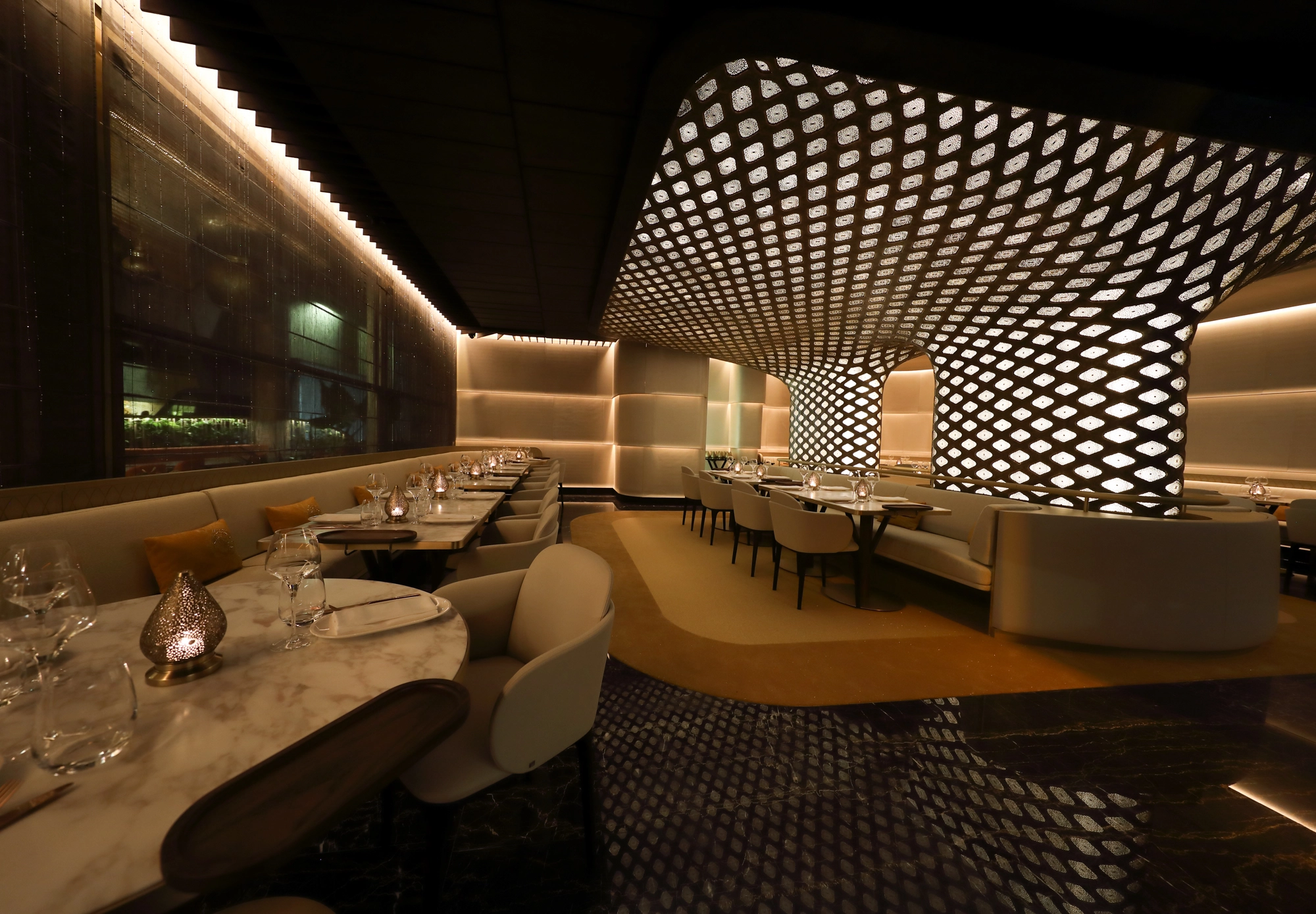 Dim restaurant where a curving perforated wall glows above a banquette and a set table.
