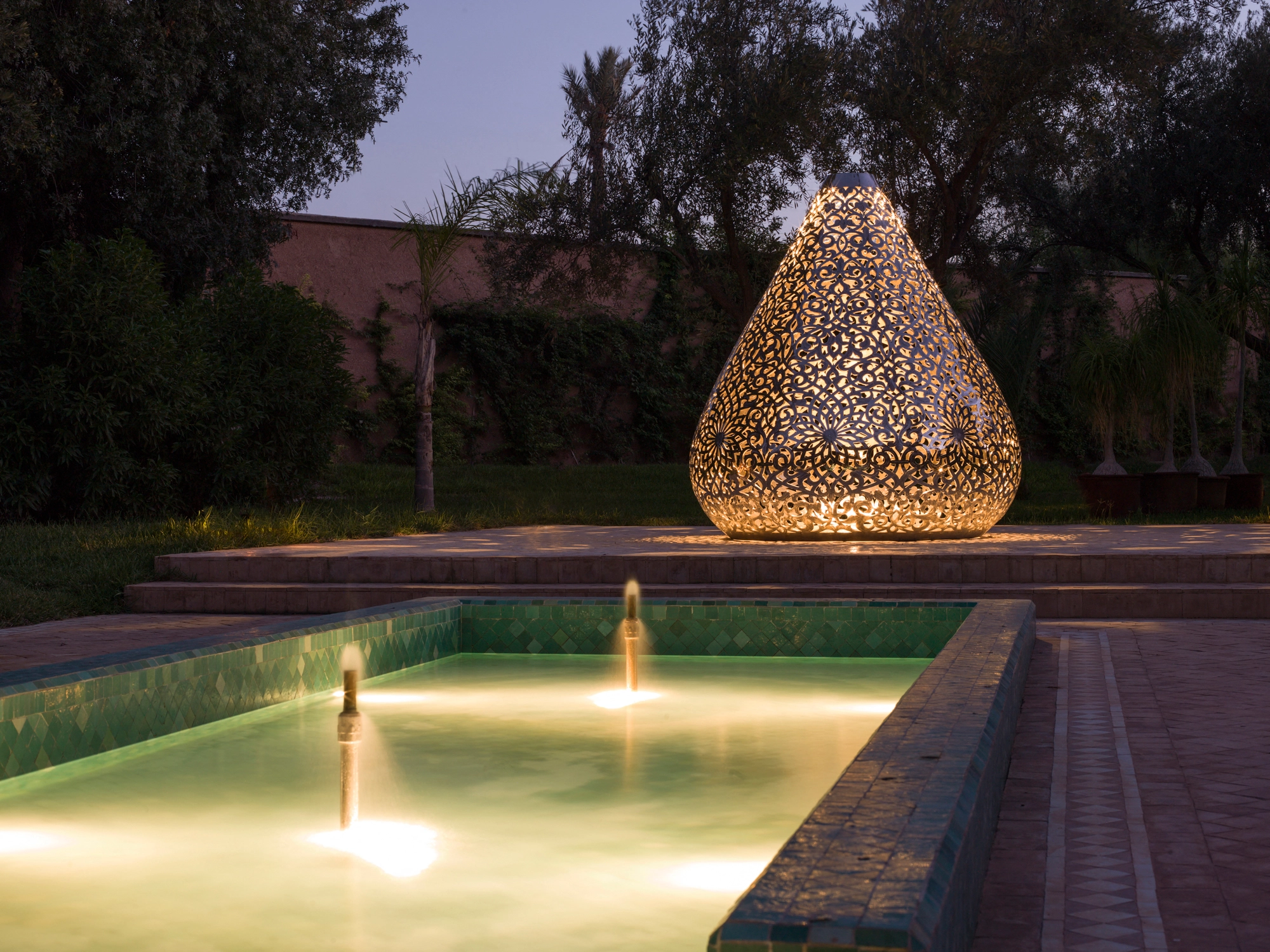 Teardrop-shaped openwork metal lotus sculpture lit at dusk beside a reflecting pool in a garden, Apollo Lotus.