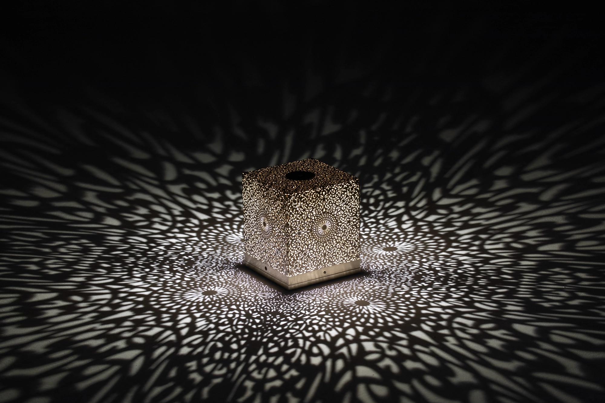 Small pierced cube table lamp glowing on a dark surface, scattering a wide starburst of light dots.