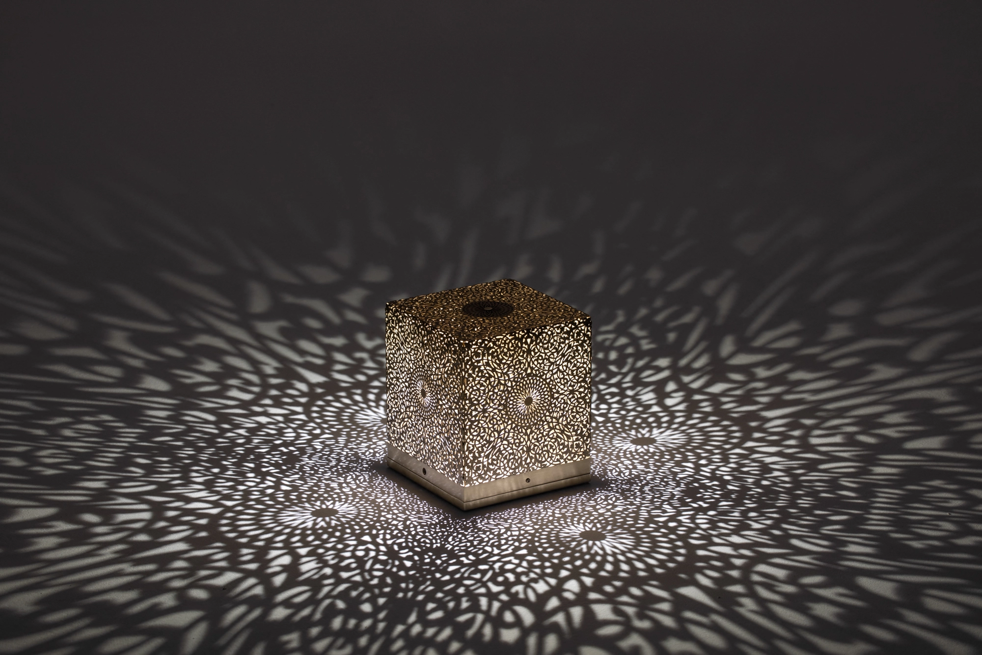 Pierced cube table lamp glowing softly, casting a halo of light dots across a dark tabletop.