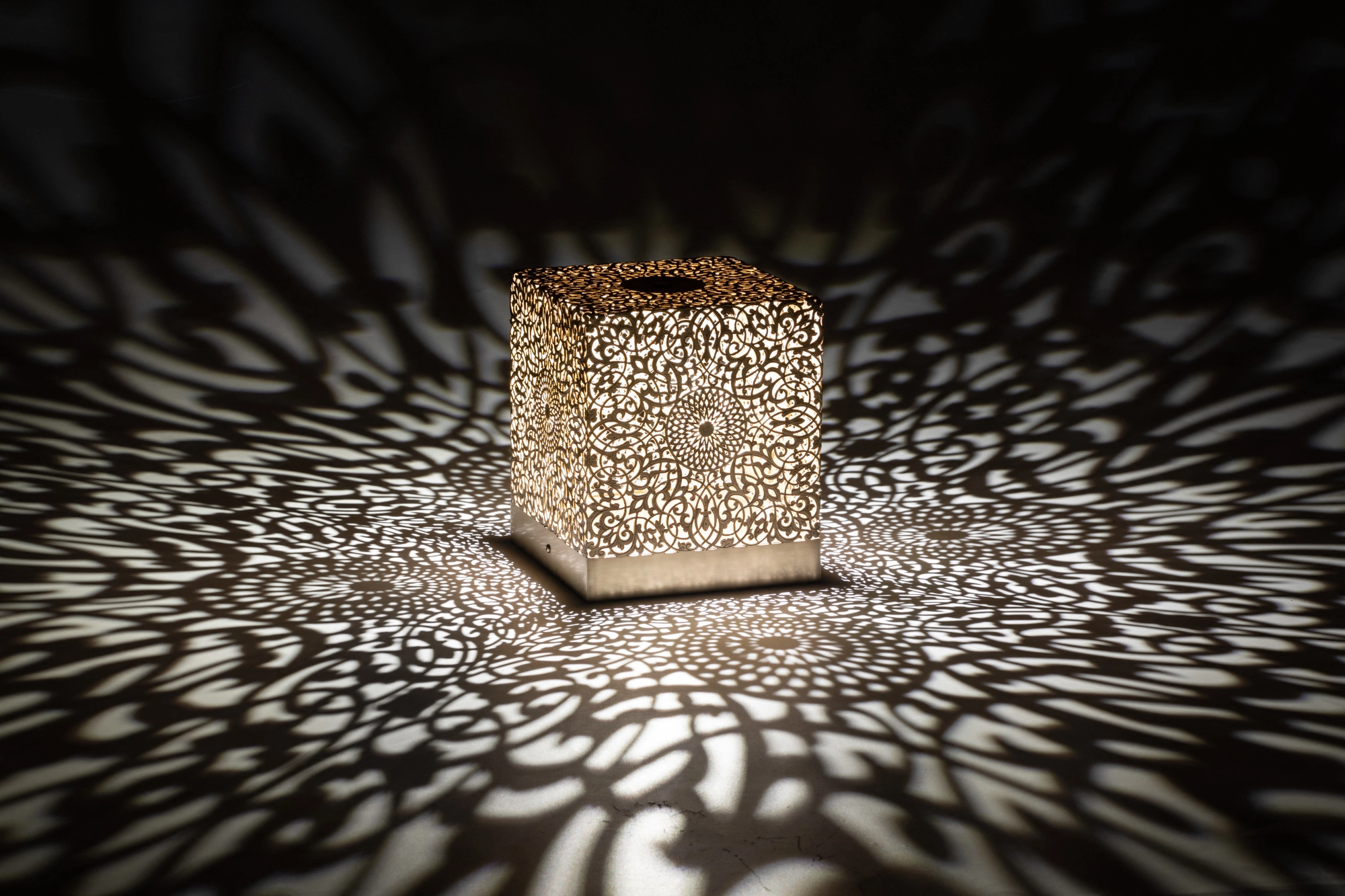 Illuminated pierced cube table lamp casting a dense scatter of light dots on a dark surface.
