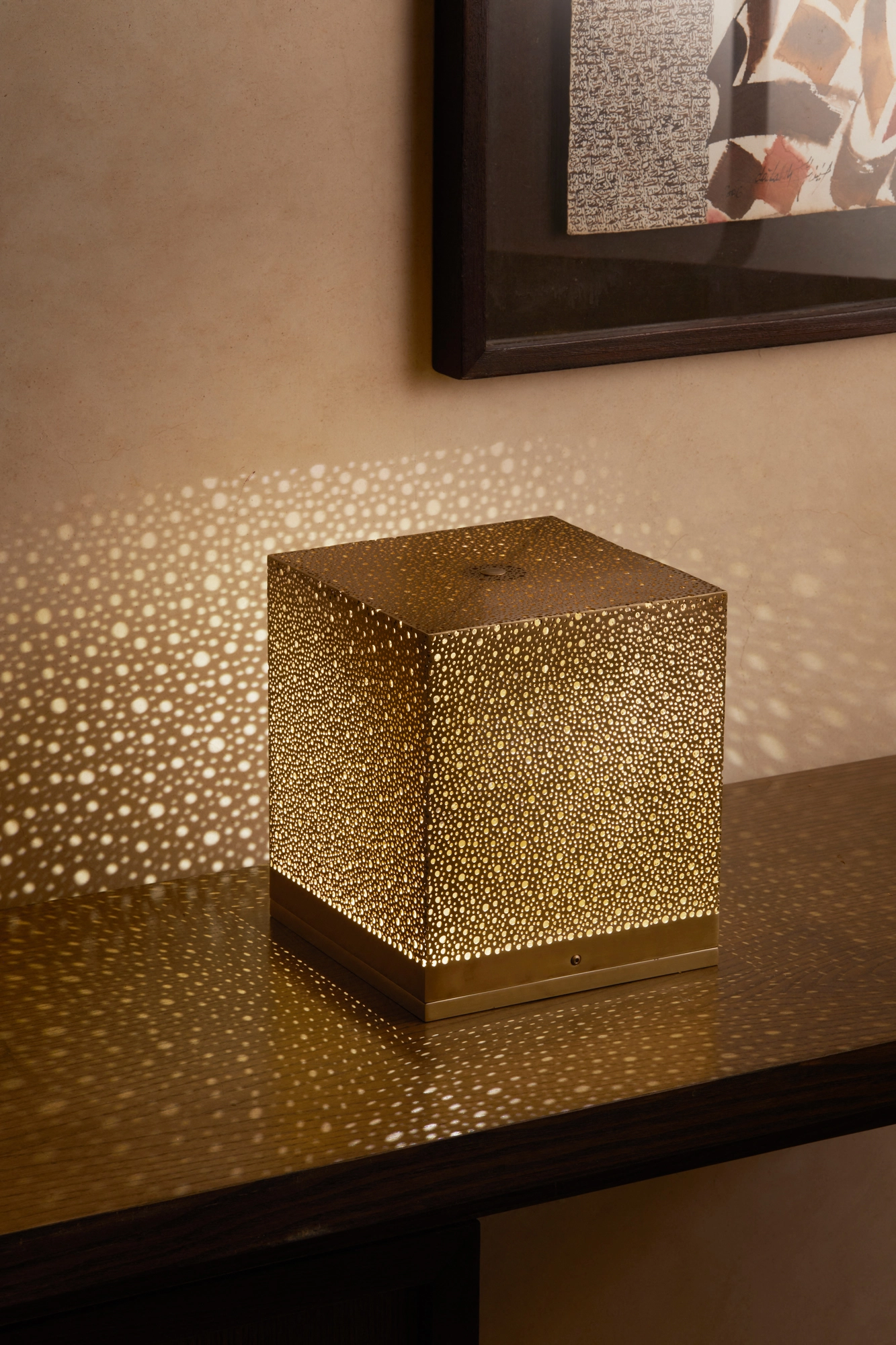 Brass pierced cube lamp glowing on a shelf beside a framed picture, scattering light dots on the wall.