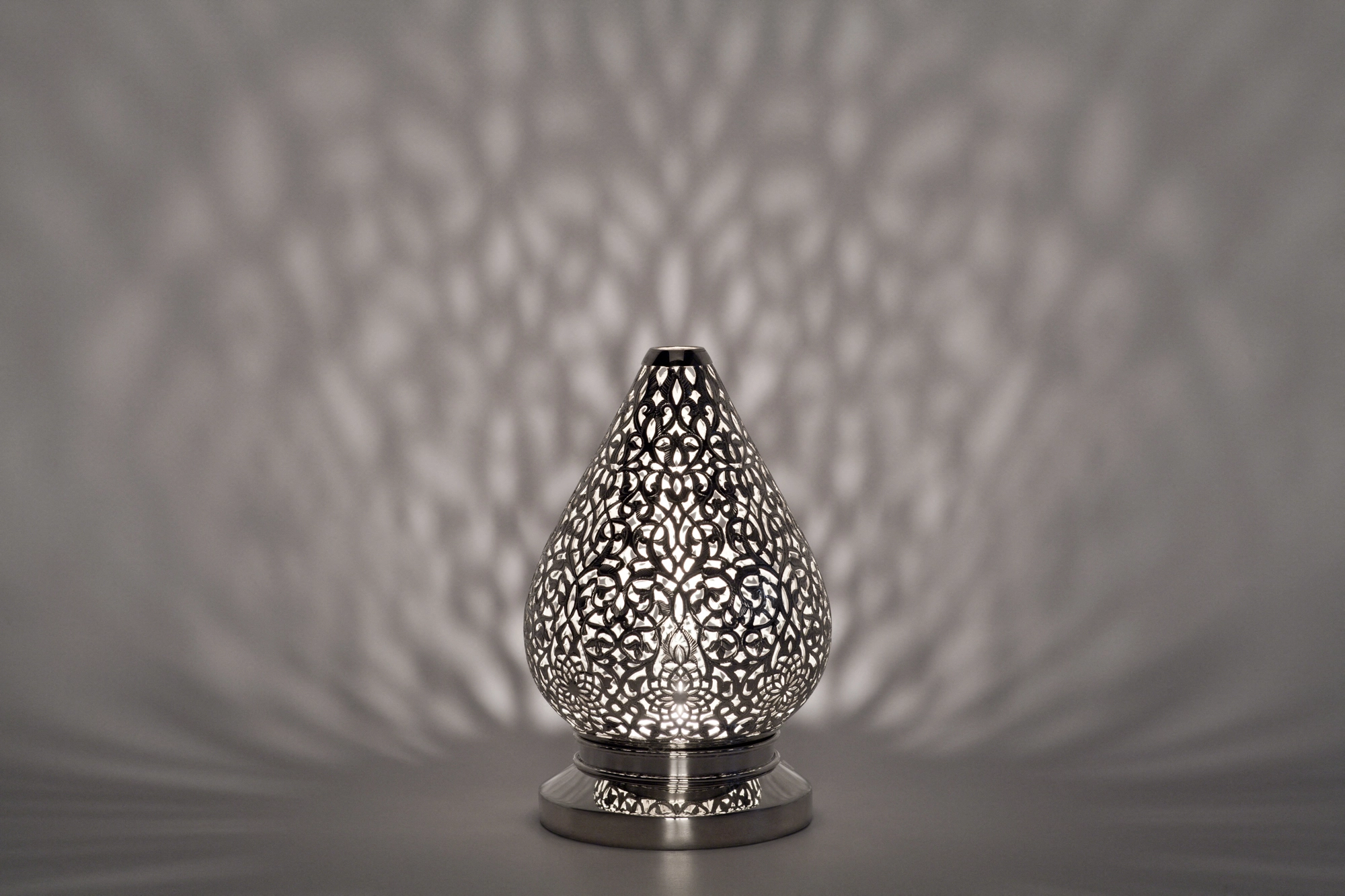 A small teardrop-shaped pierced metal candle holder casting soft dappled light against a grey background.
