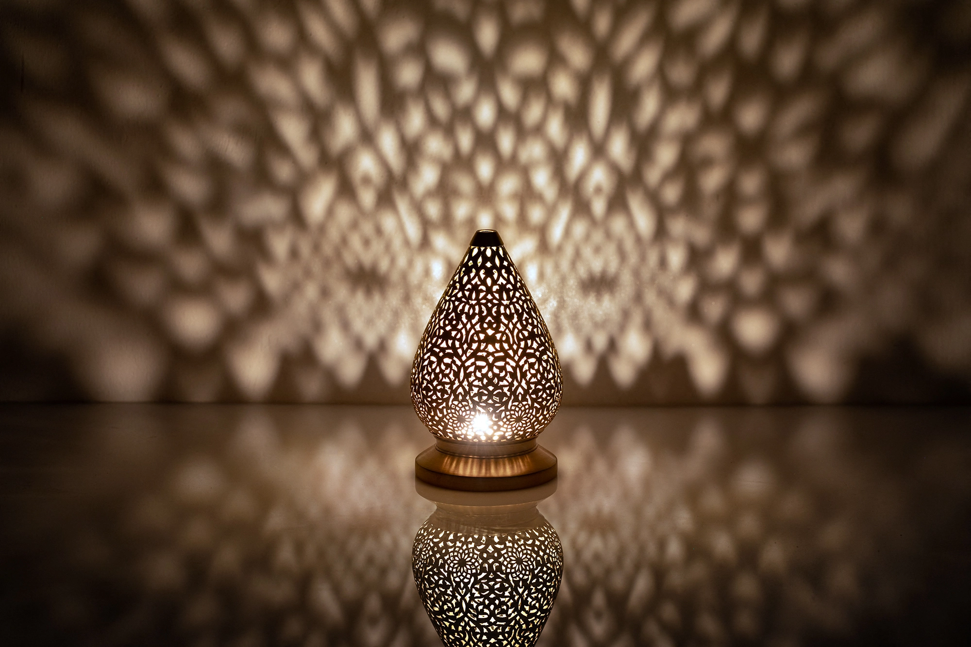 A small teardrop-shaped pierced metal candle holder glowing warmly above its reflection on a dark surface.