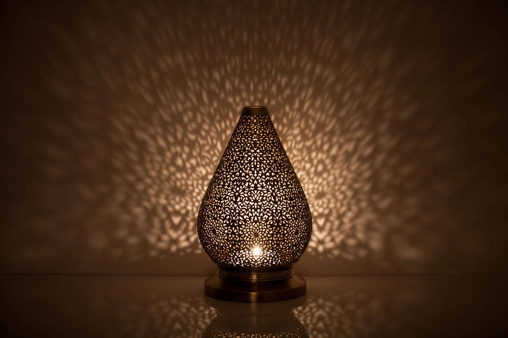 A teardrop-shaped pierced metal candle holder glowing warmly and scattering dappled light on a dark background.