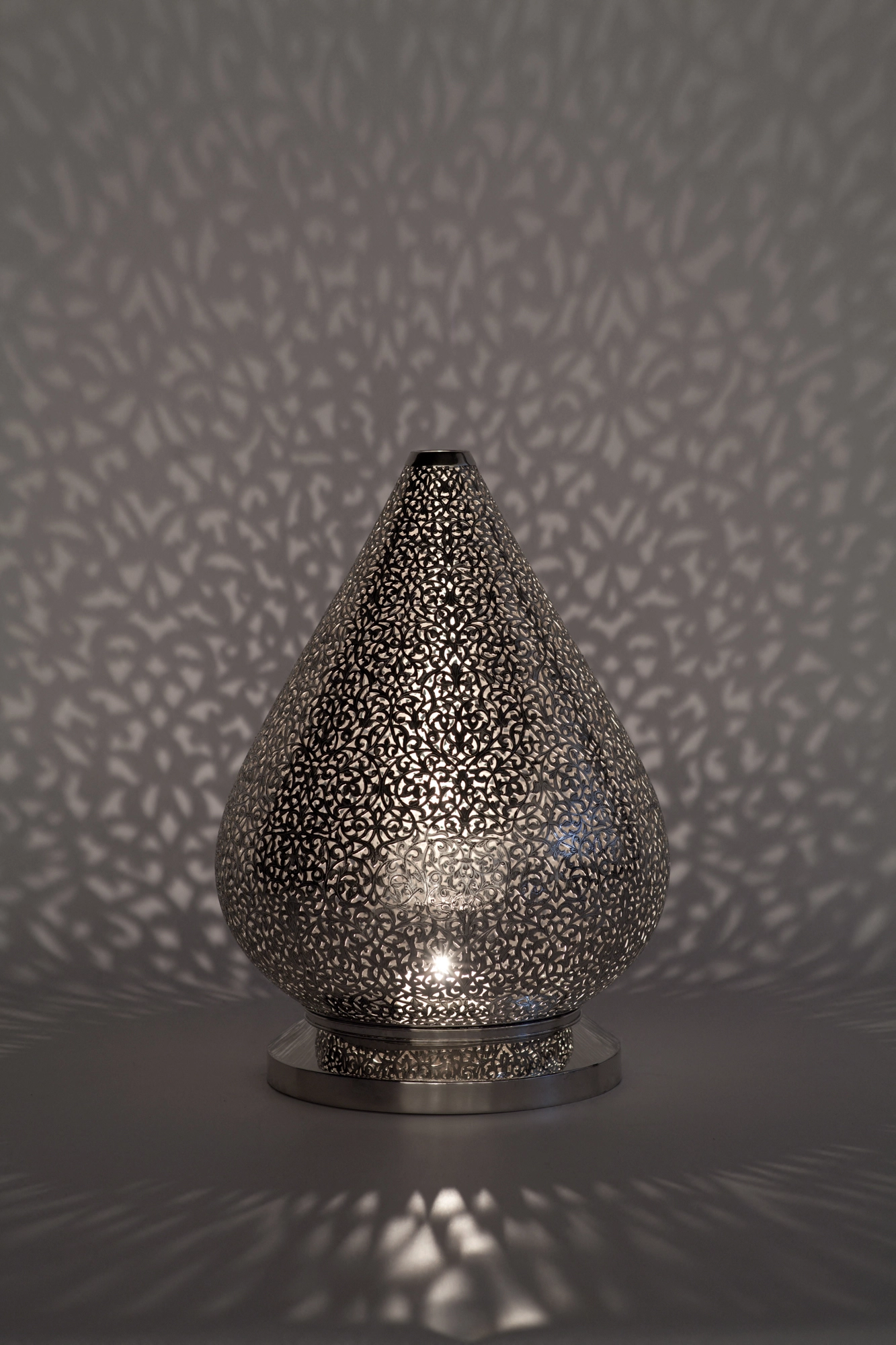 A teardrop-shaped pierced metal candle holder casting soft dappled light against a grey background.