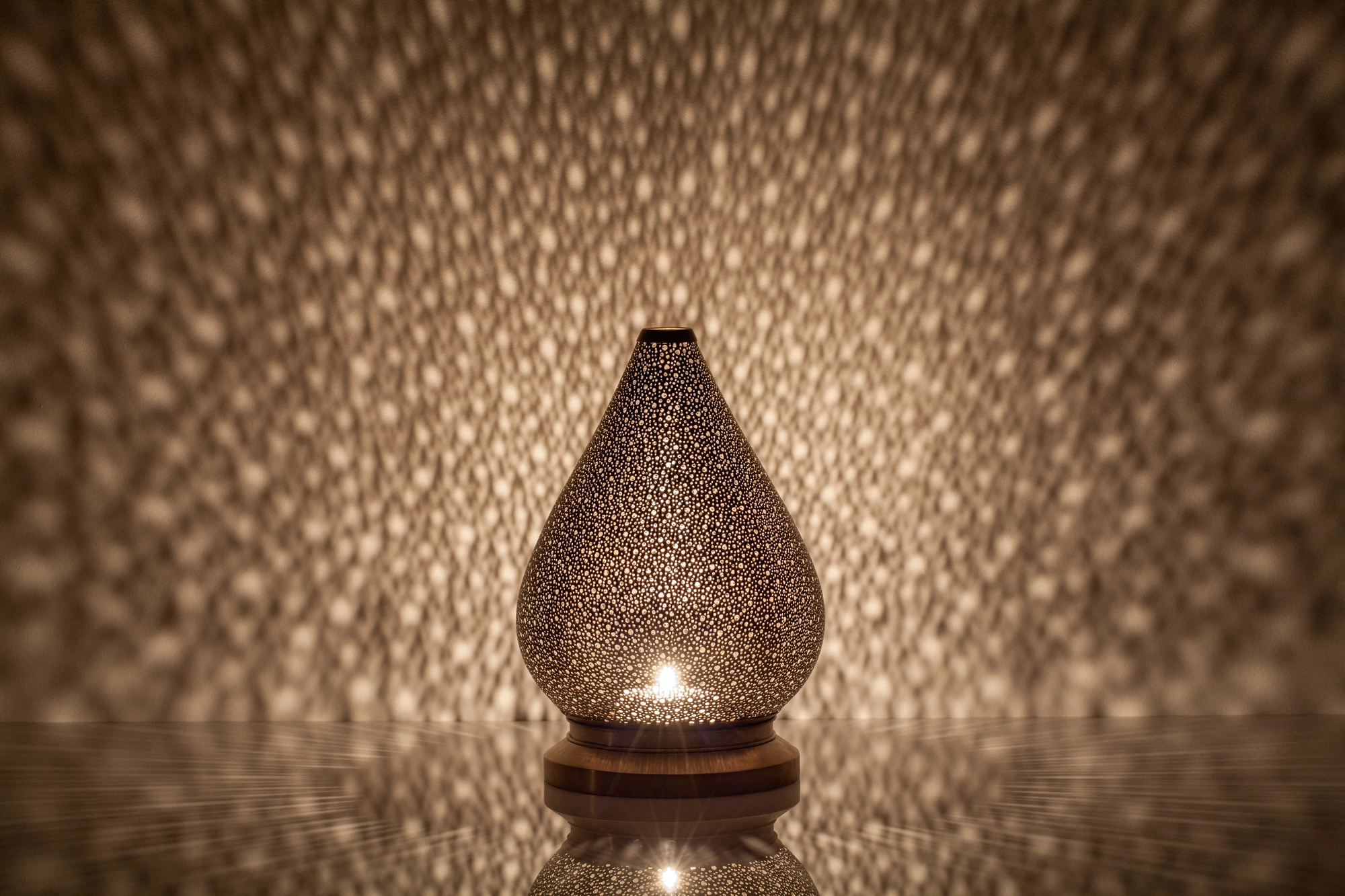 A teardrop-shaped pierced metal candle holder glowing warm amber above its reflection on a dark surface.