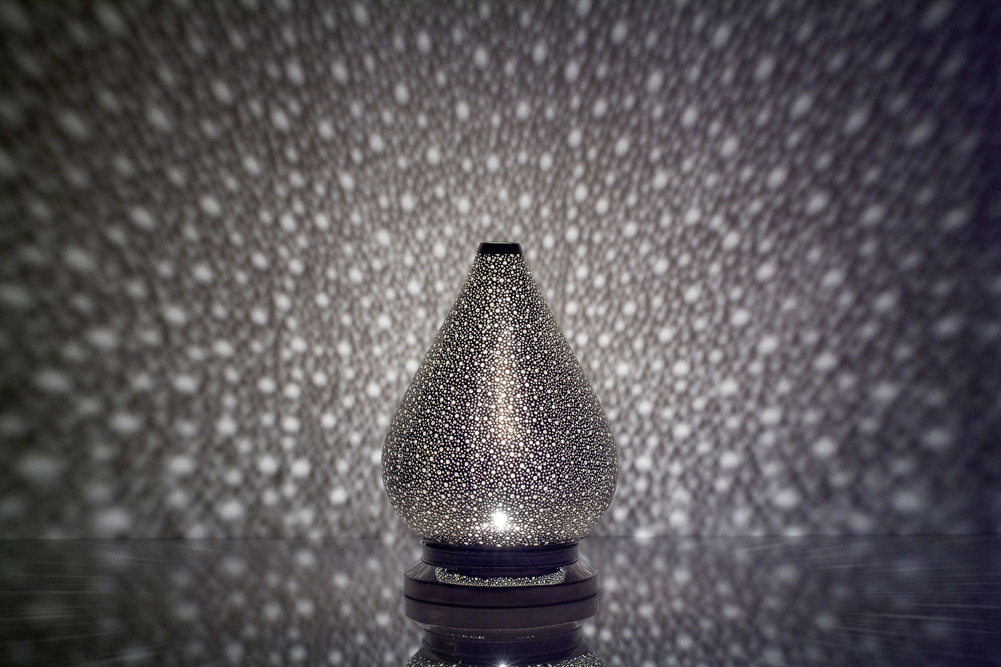 A tapered teardrop pierced metal candle holder casting fine dappled light against a soft grey background.