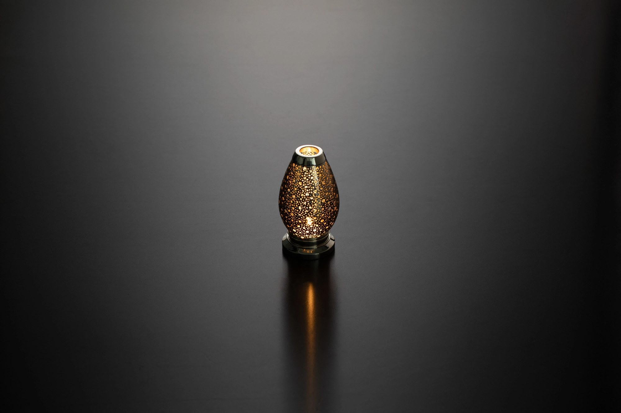 A small egg-shaped pierced metal candle holder glowing softly above its reflection on a dark surface.