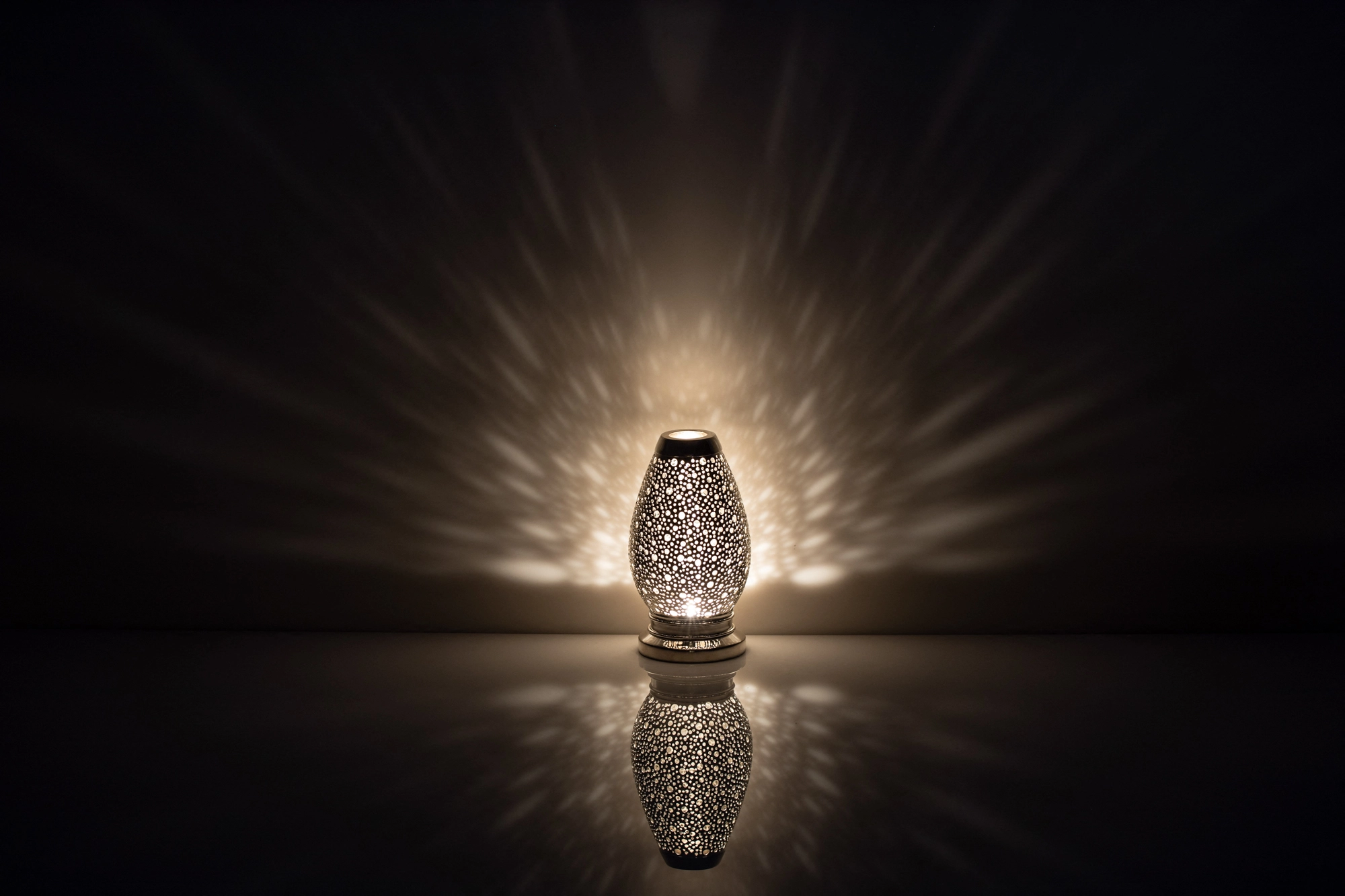 A small egg-shaped pierced metal candle holder glowing brightly above its reflection on a dark surface.