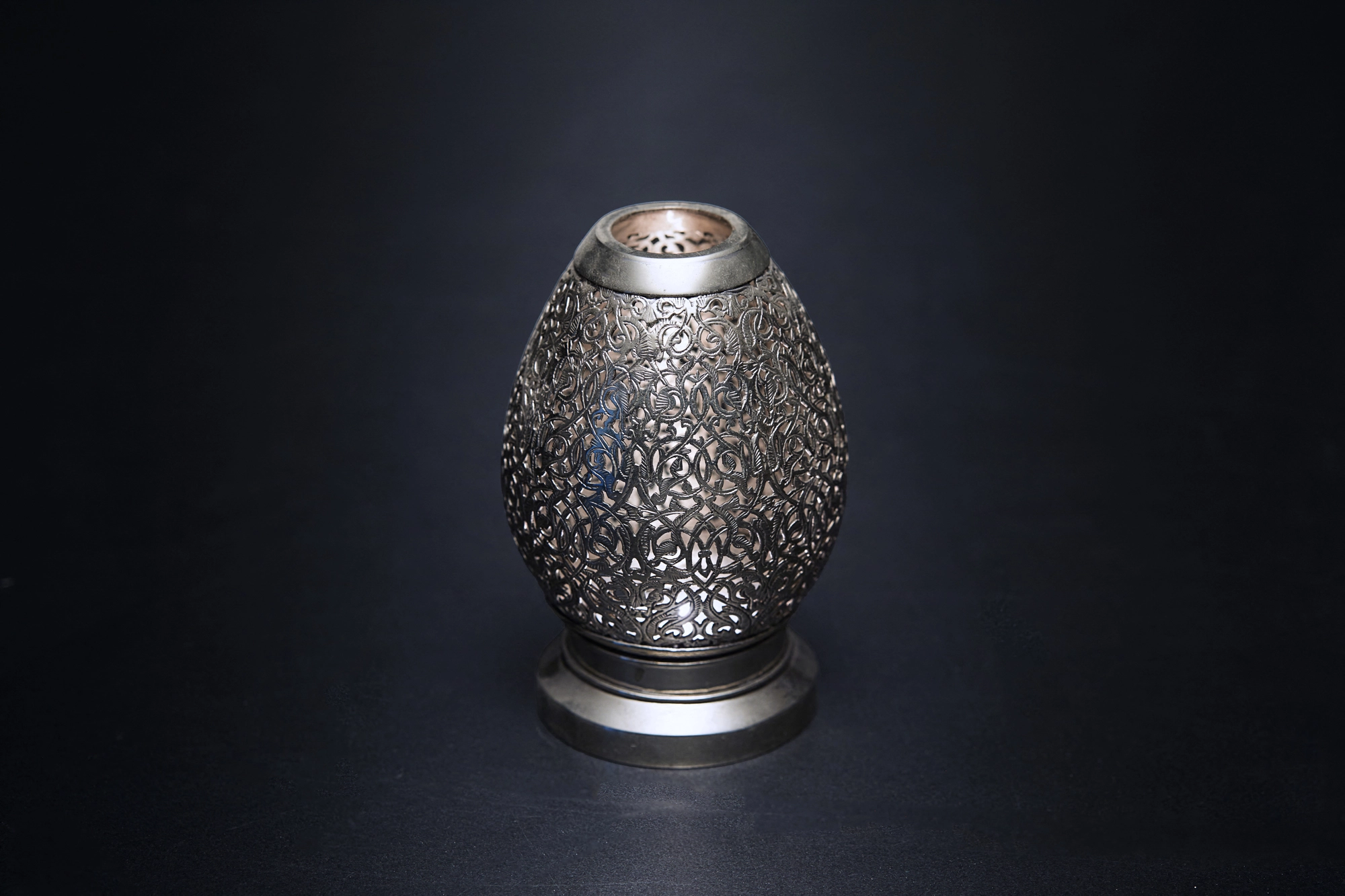 A small egg-shaped pierced metal candle holder glowing dimly against a dark background.