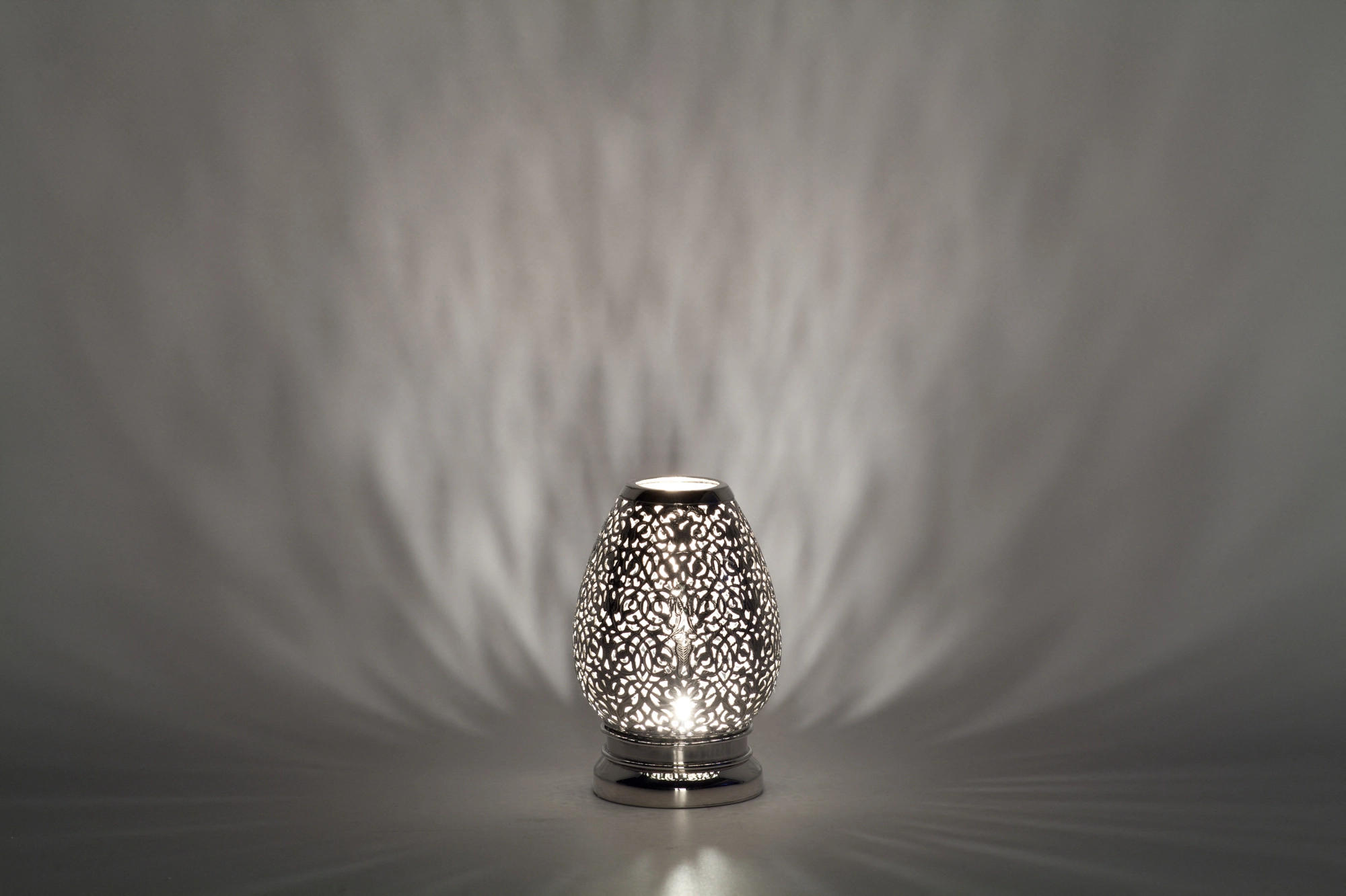 A small egg-shaped pierced metal candle holder scattering pale dappled light against a dark background.