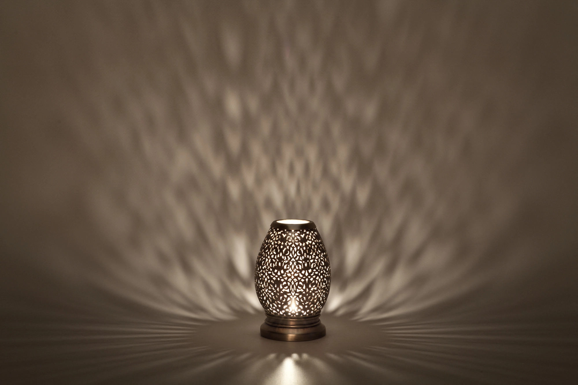 A small egg-shaped pierced metal candle holder casting warm dappled light against a dark background.
