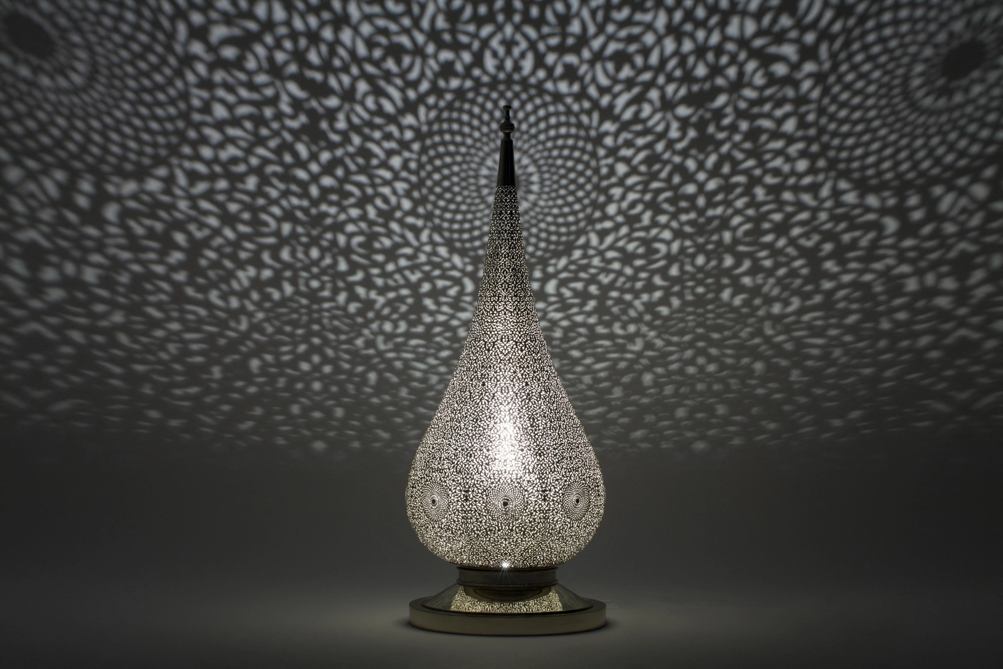 A pierced teardrop floor lamp glowing on a base, scattering a fine dotted spray of light on a grey ground.