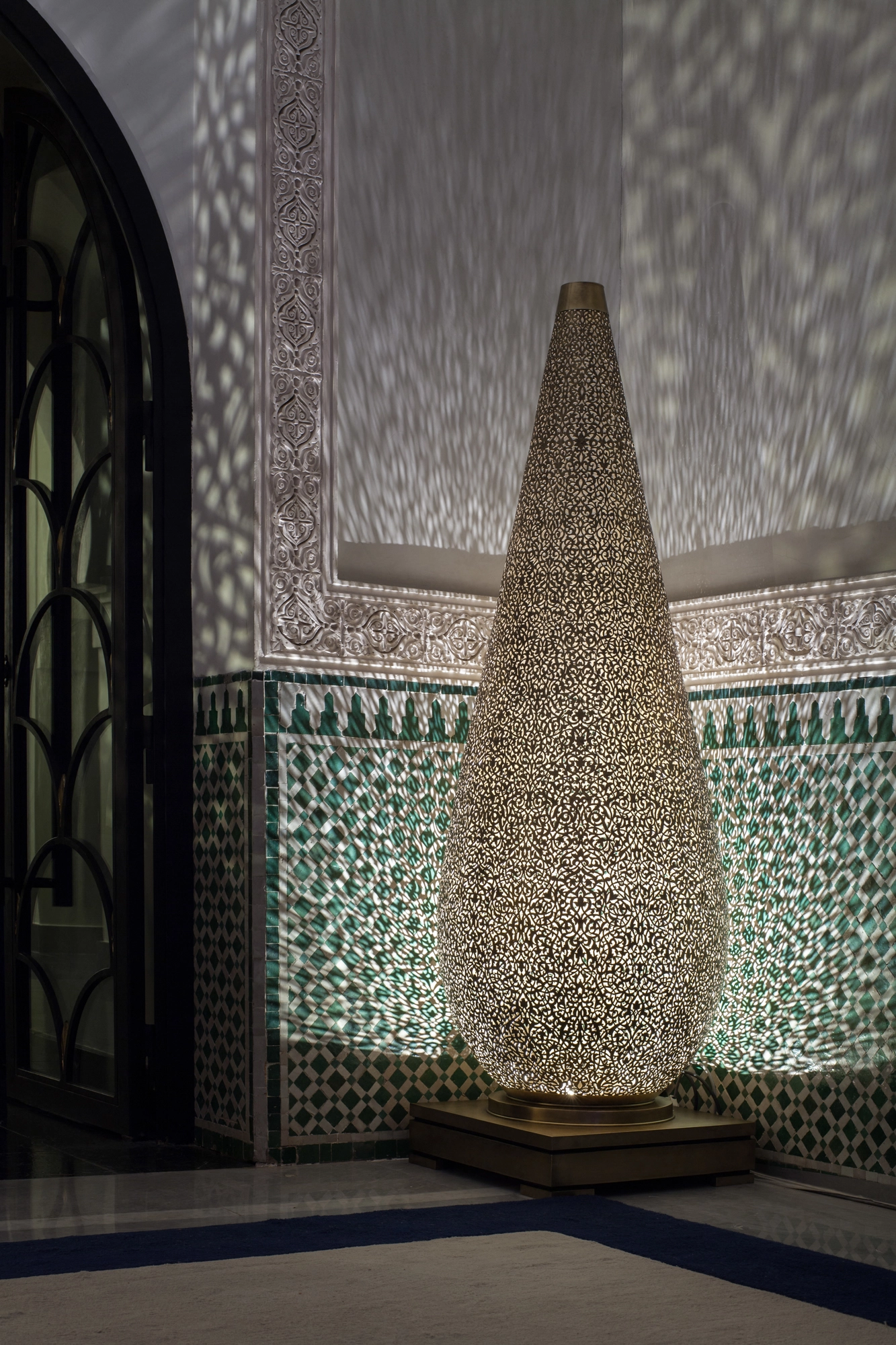 Tall pierced teardrop floor lamp glowing against a green zellige tiled wall, scattering light patterns.