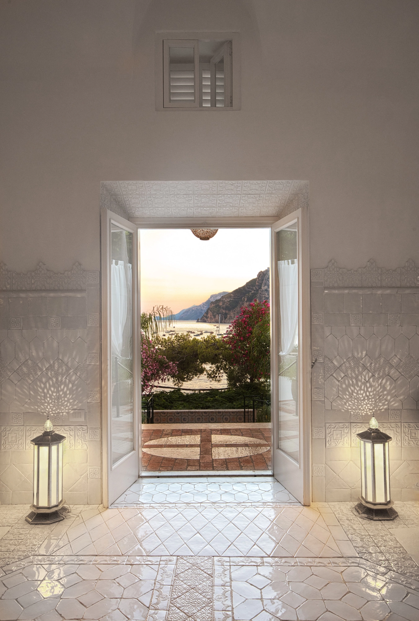 Open doorway framing a coastal sea view at dusk, flanked by two lantern floor lamps on a tiled floor.