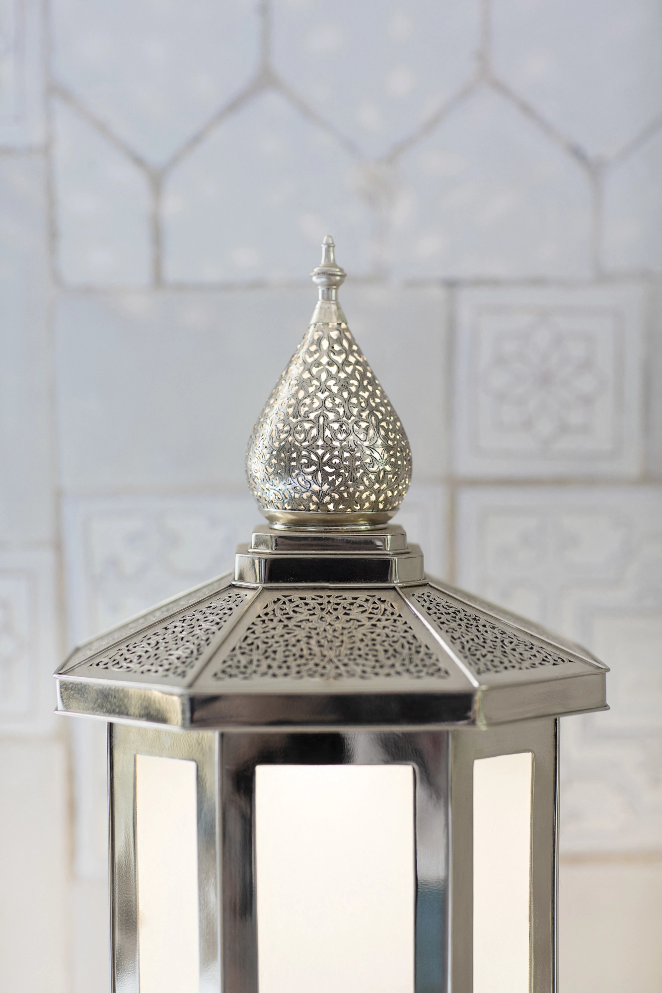 Pierced silver lantern with an onion-dome top standing against a white tiled wall.